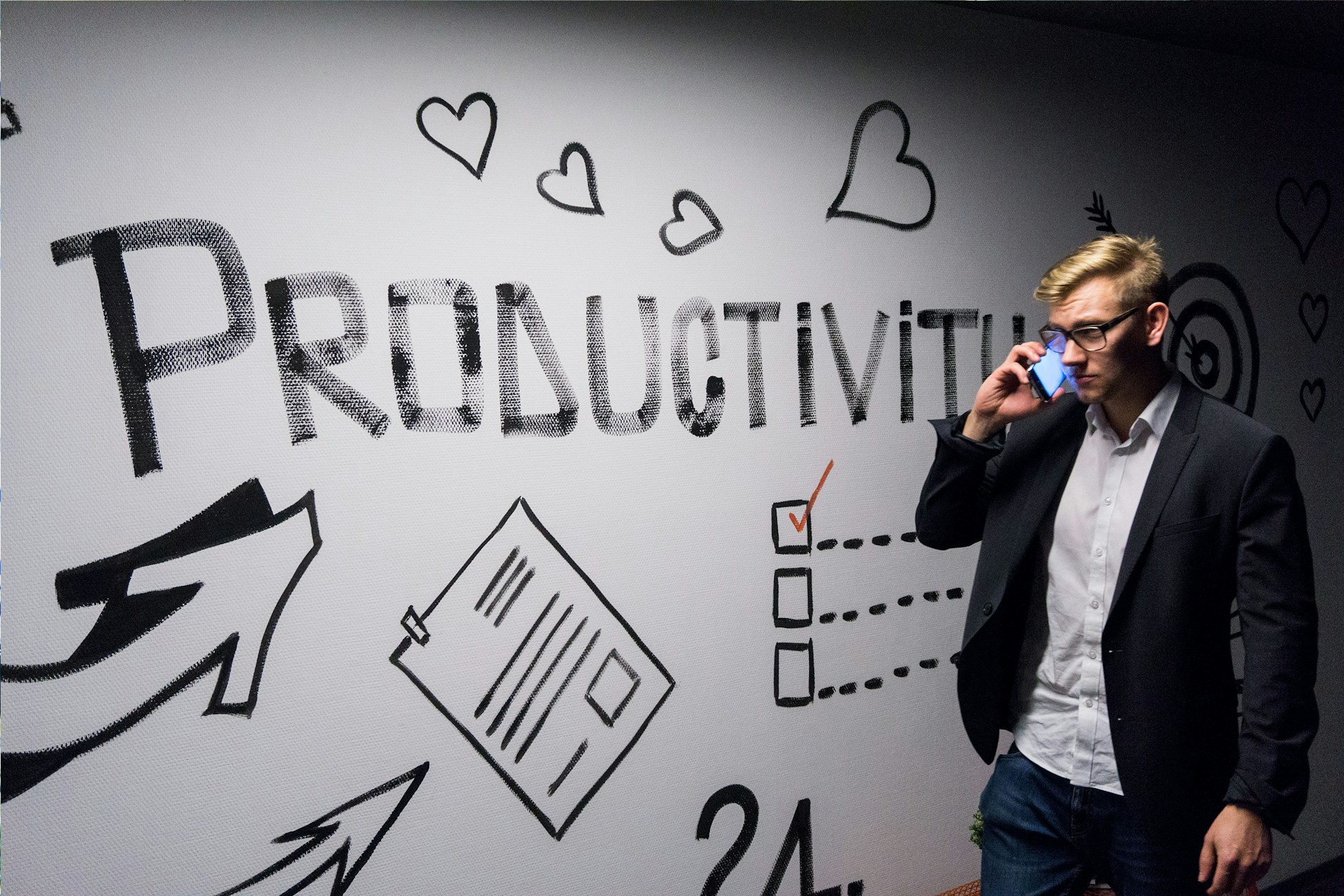 Colour photograph of man walking in front of a white wall with the word "productivity" on it while speaking on the phone, by Andreas Klassen on unsplash.
