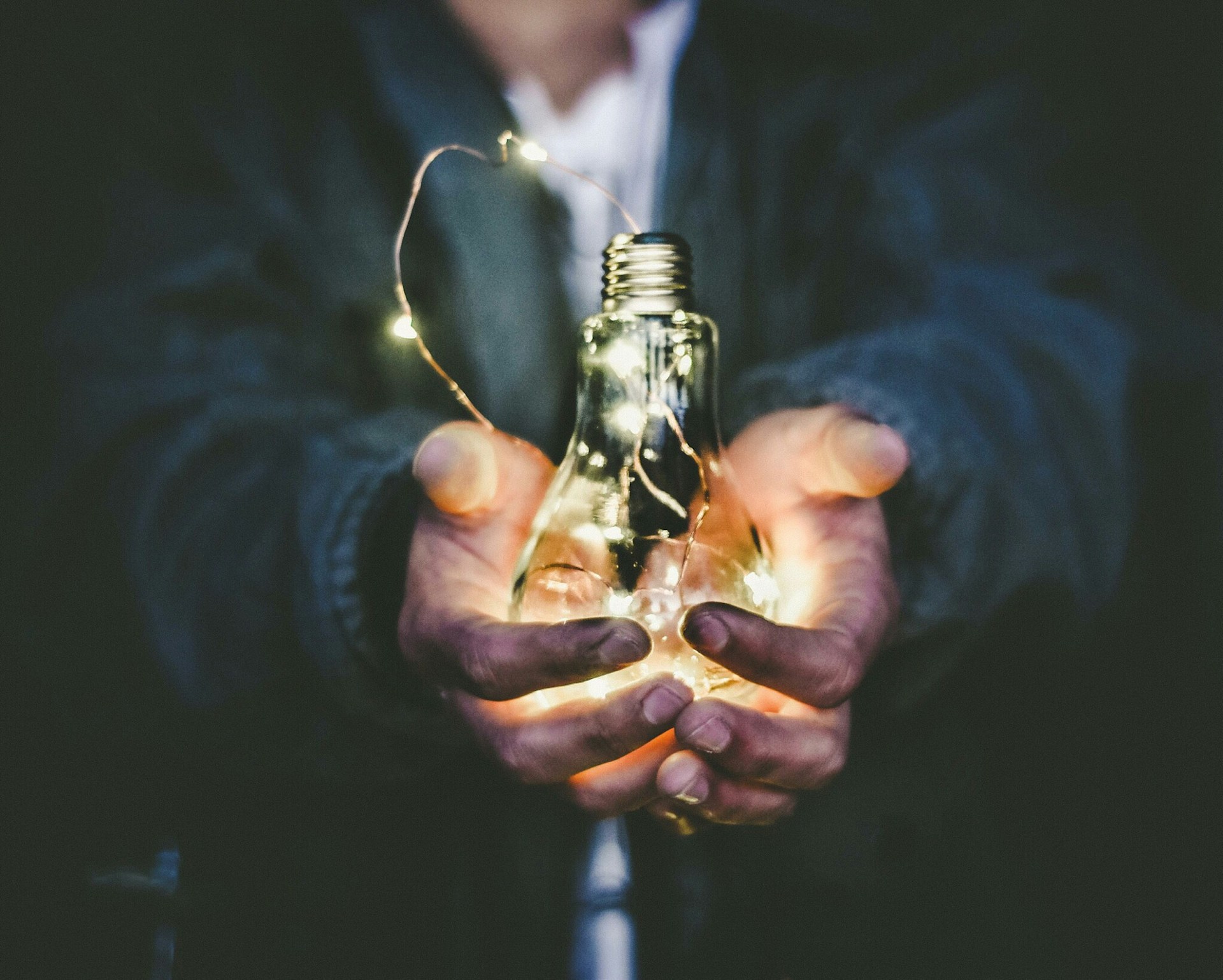 Colour photograph of a man holding a lit light bulb in his hands, close-up of the hands and light bulb by Riccardo Annandale on unsplash