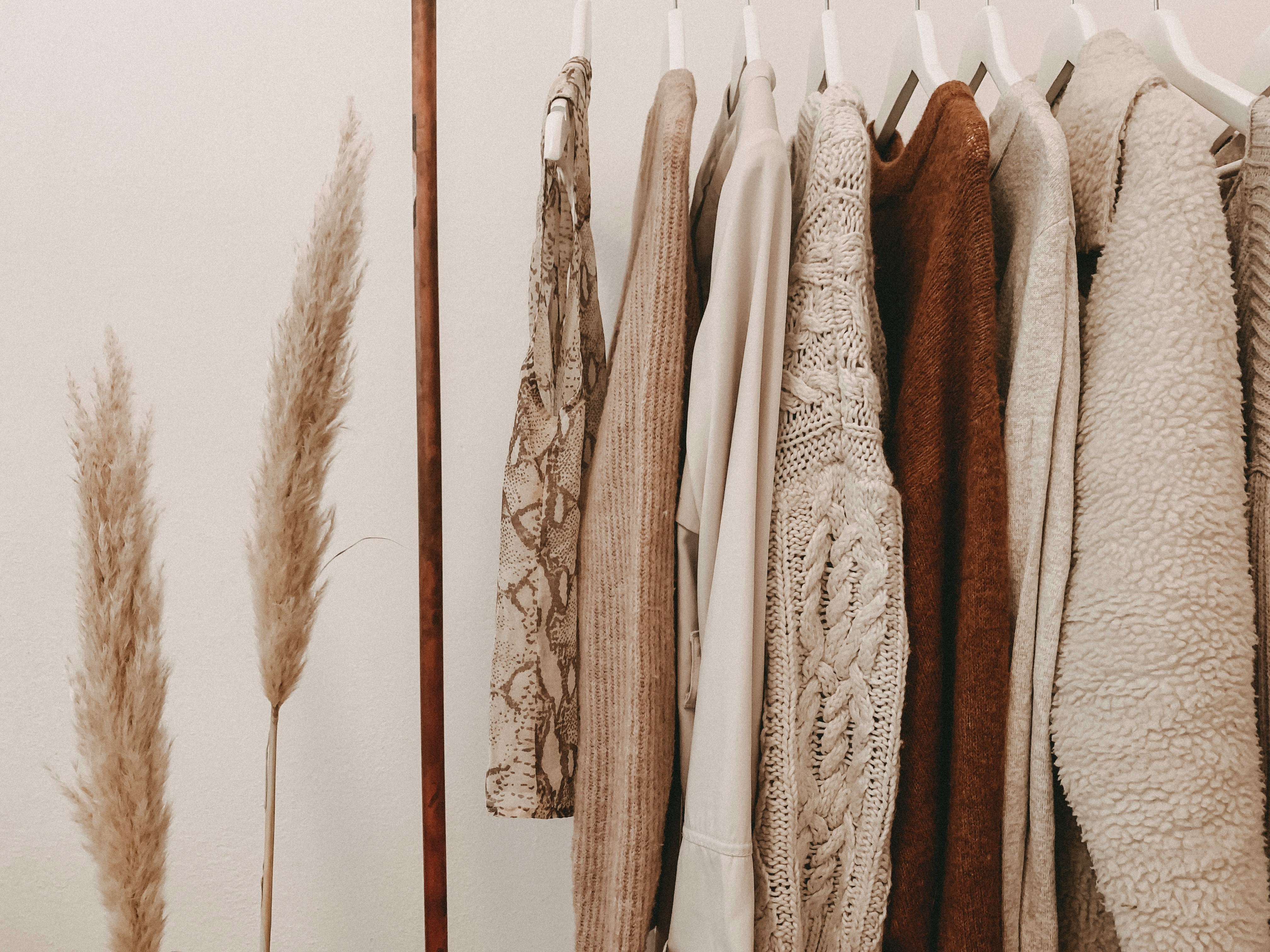 Neutral-toned sweaters, coats, and tops hanging on a clothing rack beside dried pampas grass.