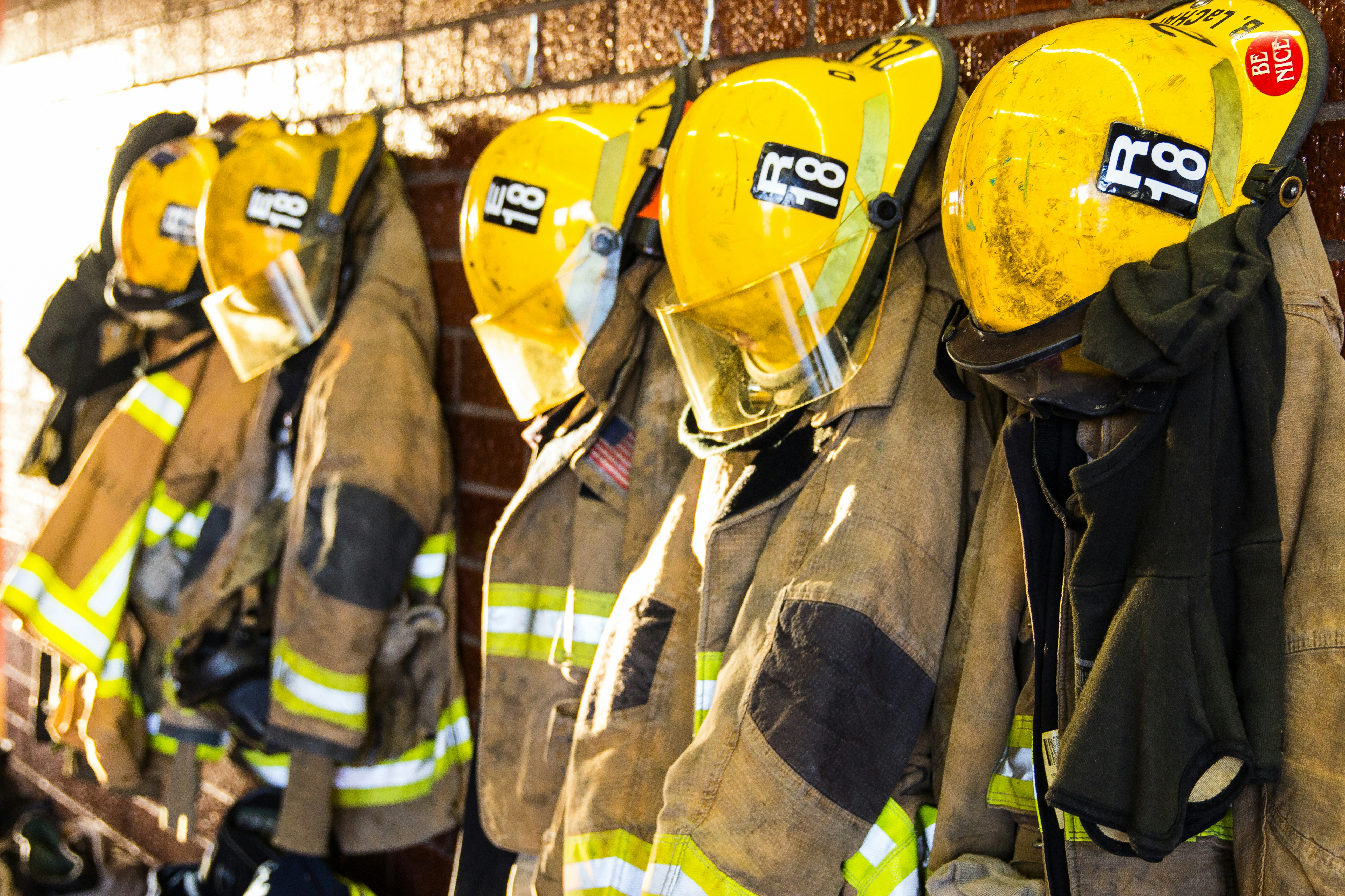 Several yellow firefighter helmets and protective turnout jackets hang on hooks along a brick wall in a fire station.