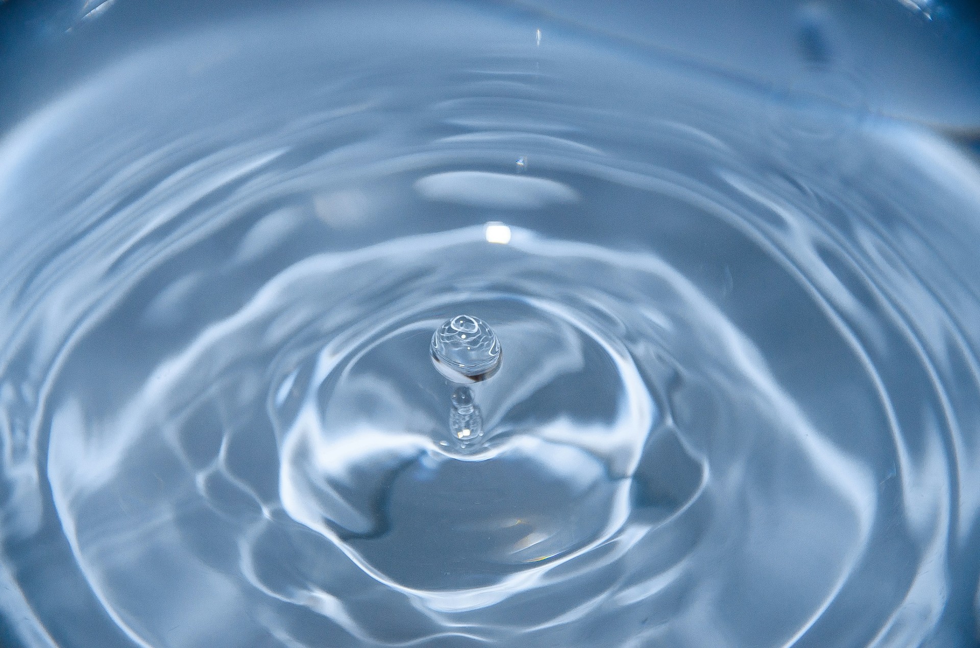 A photo presenting a droplet rebounding from a rippling blue water surface, captured in macro detail.