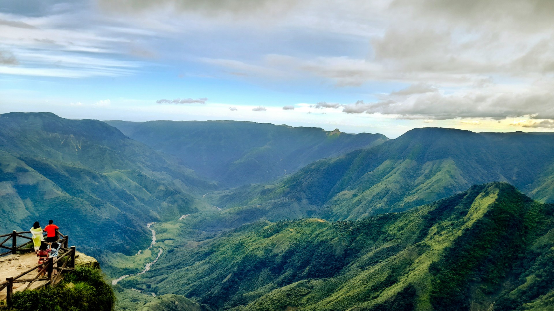 Photo presenting visitors at a lookout point overlooking a lush green river valley in the mountains.