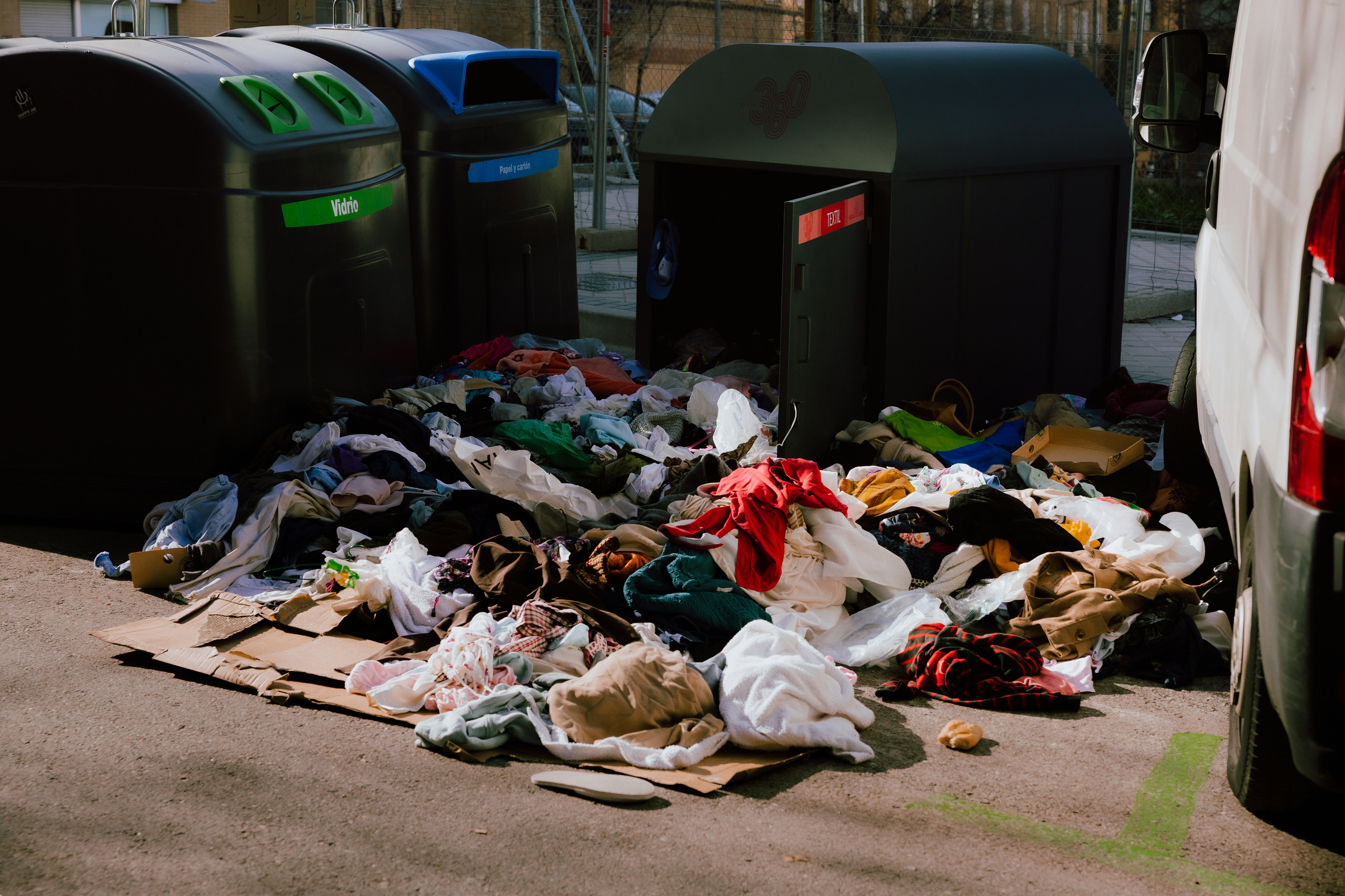 A photo presenting a pile of discarded clothing spilling out beside recycling bins on a street.