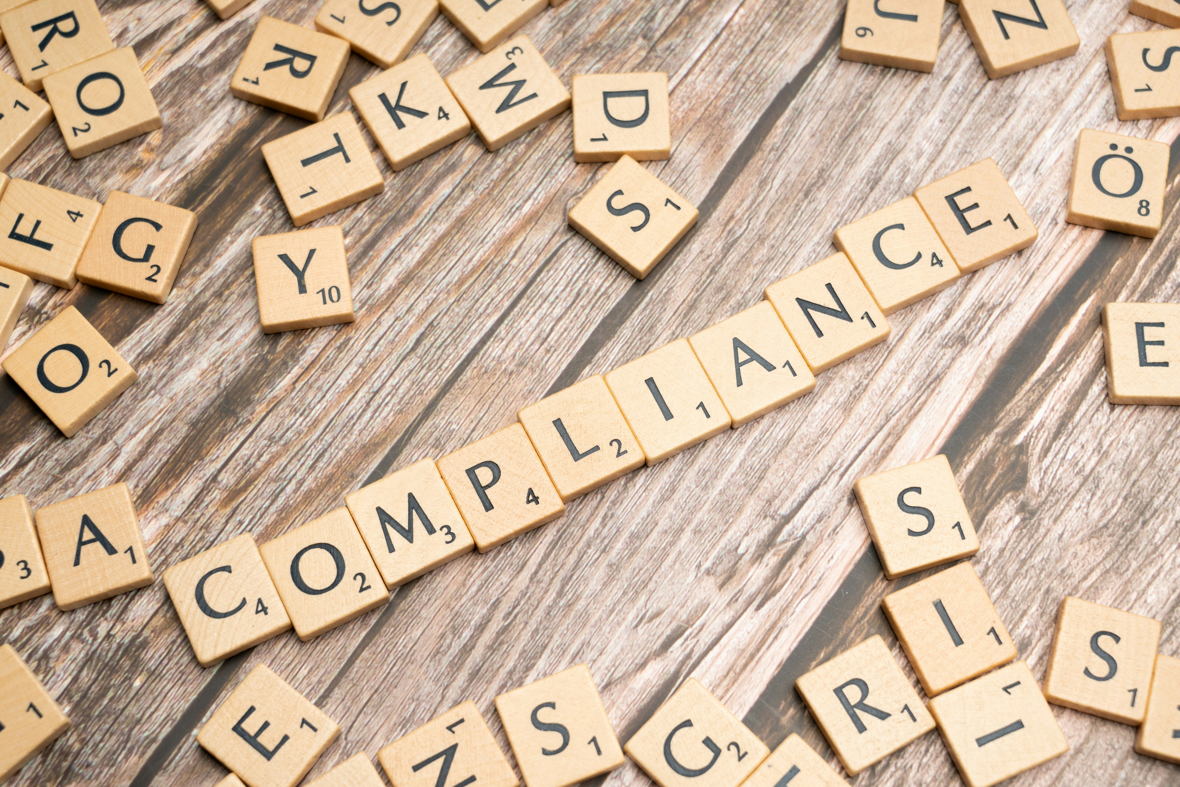 Photo presenting wooden letter tiles arranged on a rustic wooden surface, with the word “COMPLIANCE”
