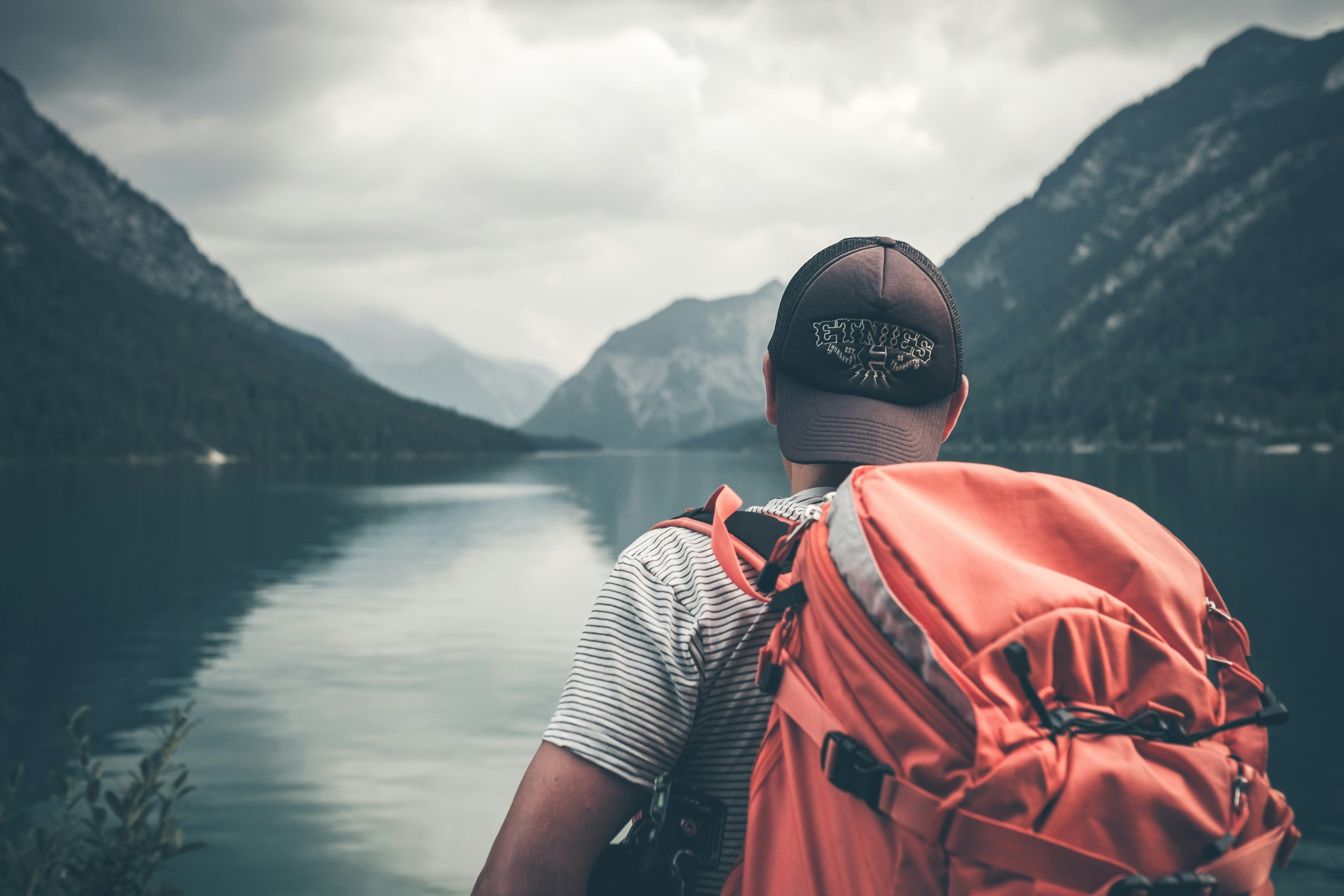 The picture presents a traveler with a backpack facing a calm mountain lake.
