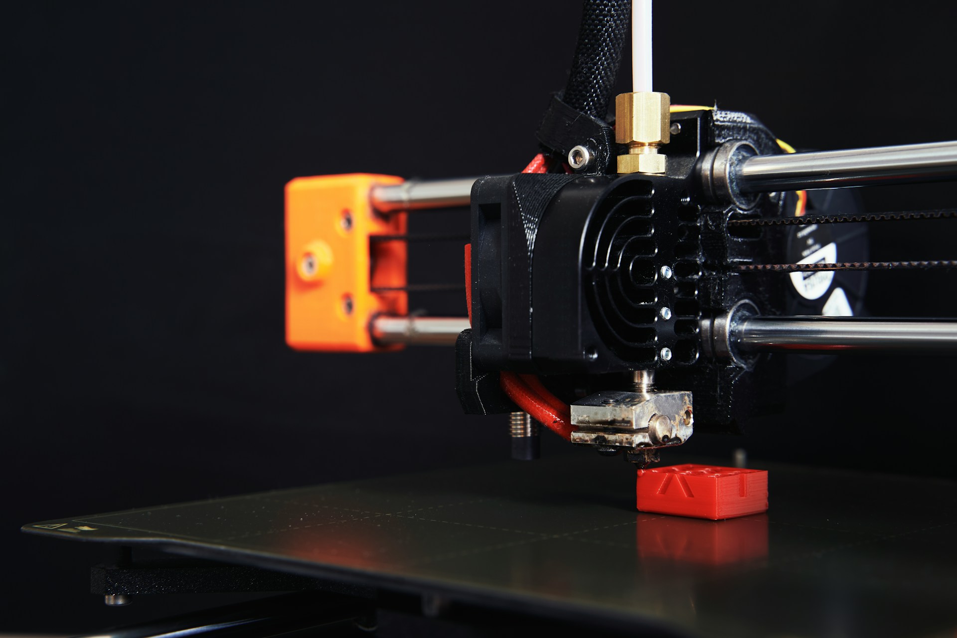 The photo presents a close-up of a 3D printer producing a small plastic component, illustrating additive manufacturing.
