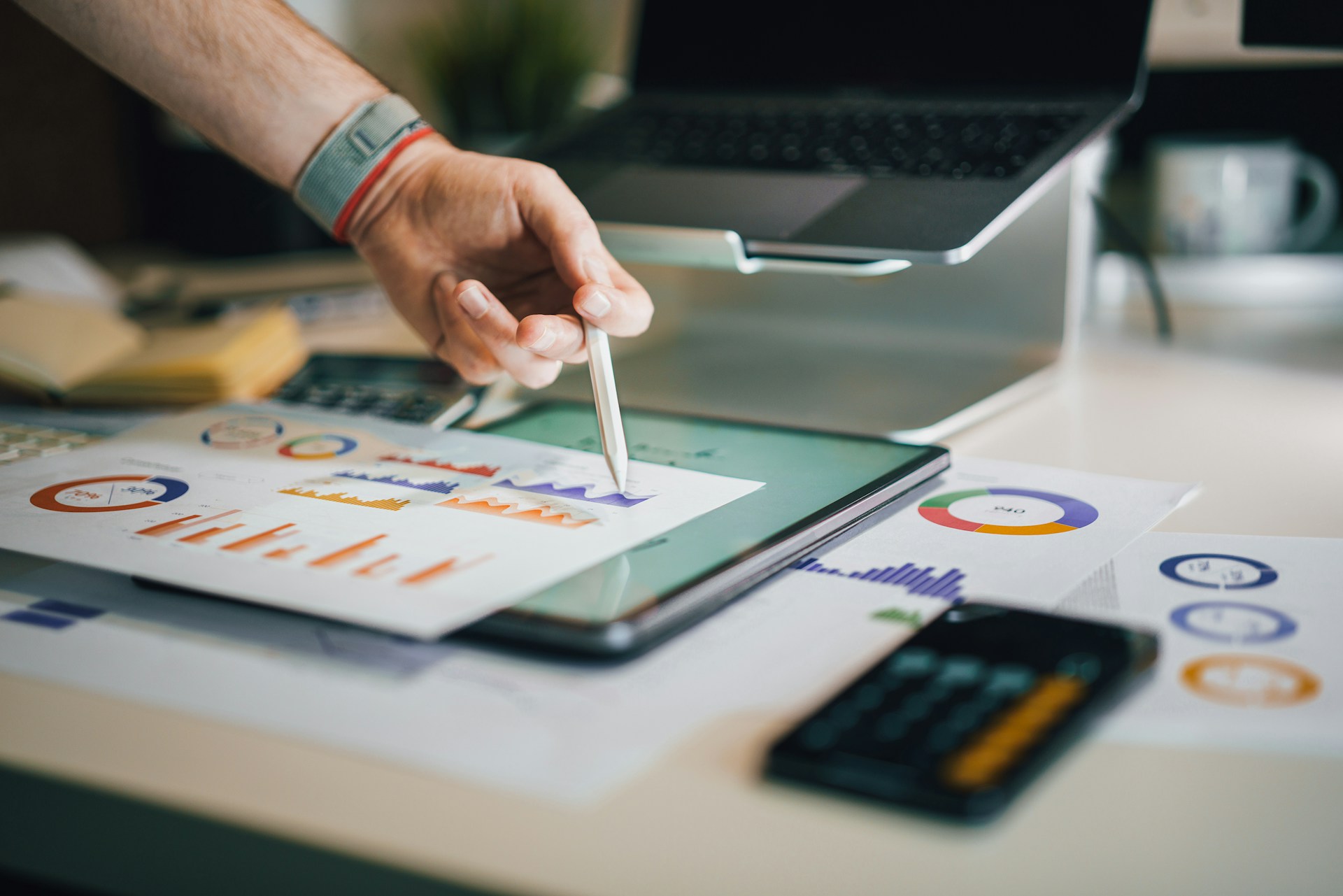 The photo presents a person analysing charts and financial data with a tablet, laptop and calculator.