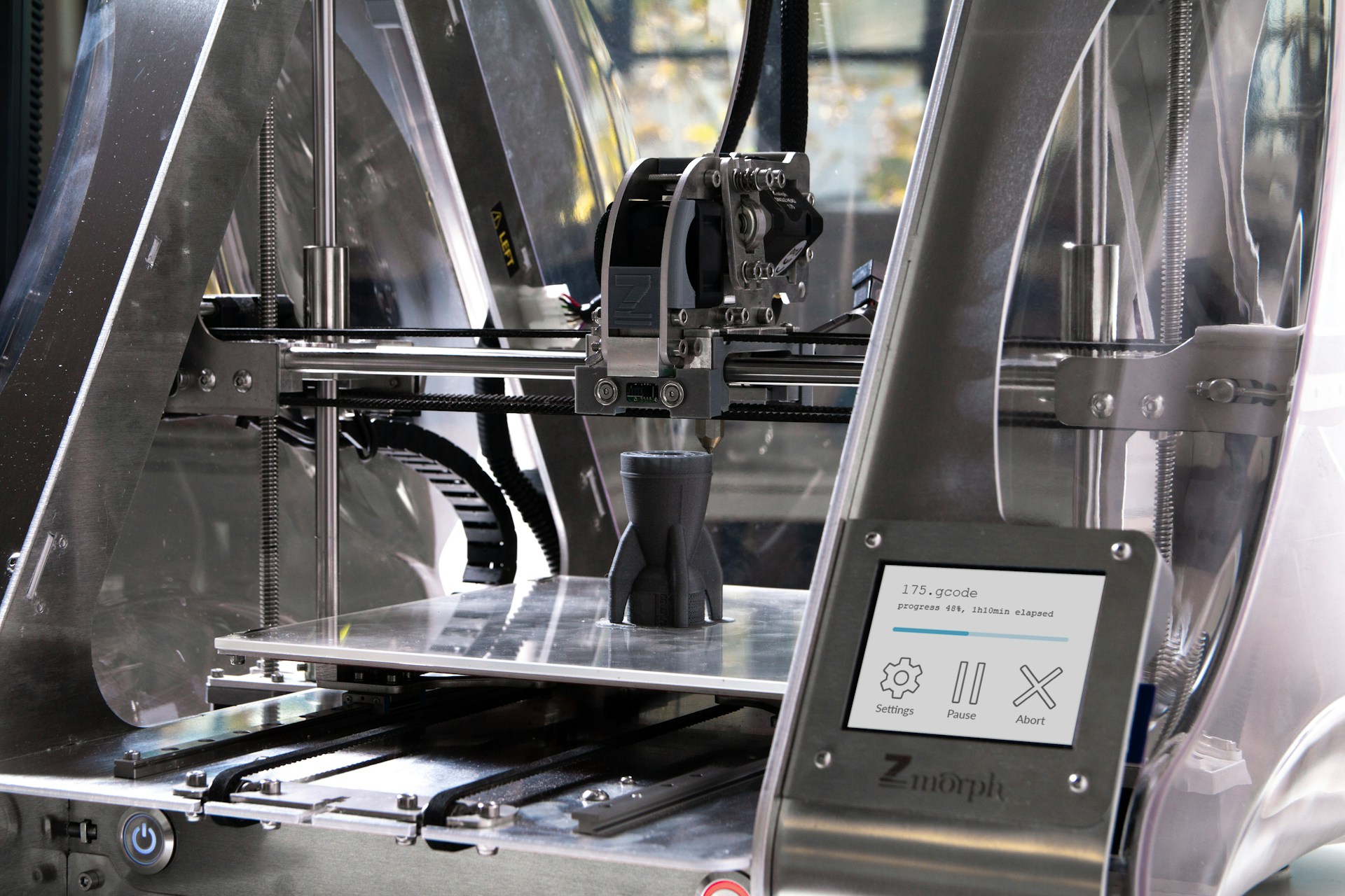 The photo presents a 3D printer producing a plastic component, illustrating additive manufacturing.