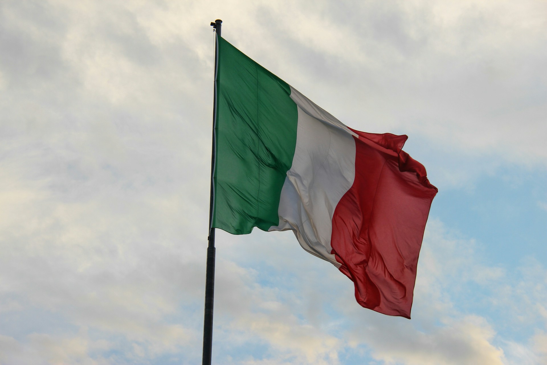The photo presents the Italian flag waving on a flagpole against a partly cloudy sky.