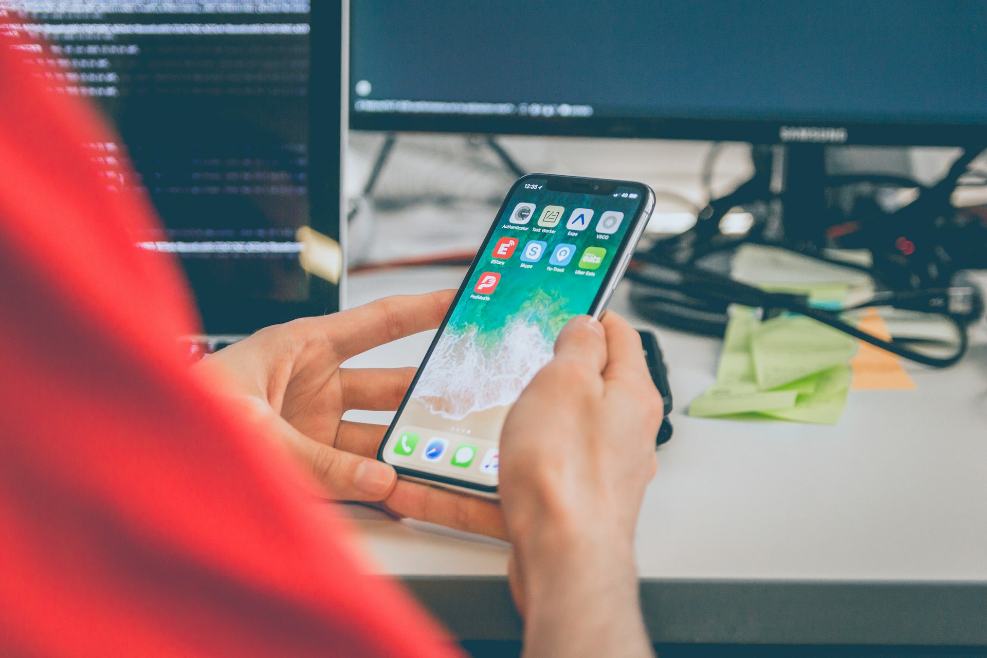 The photo presents a person holding a smartphone displaying app icons at a desk, with computer monitors showing code in the background.