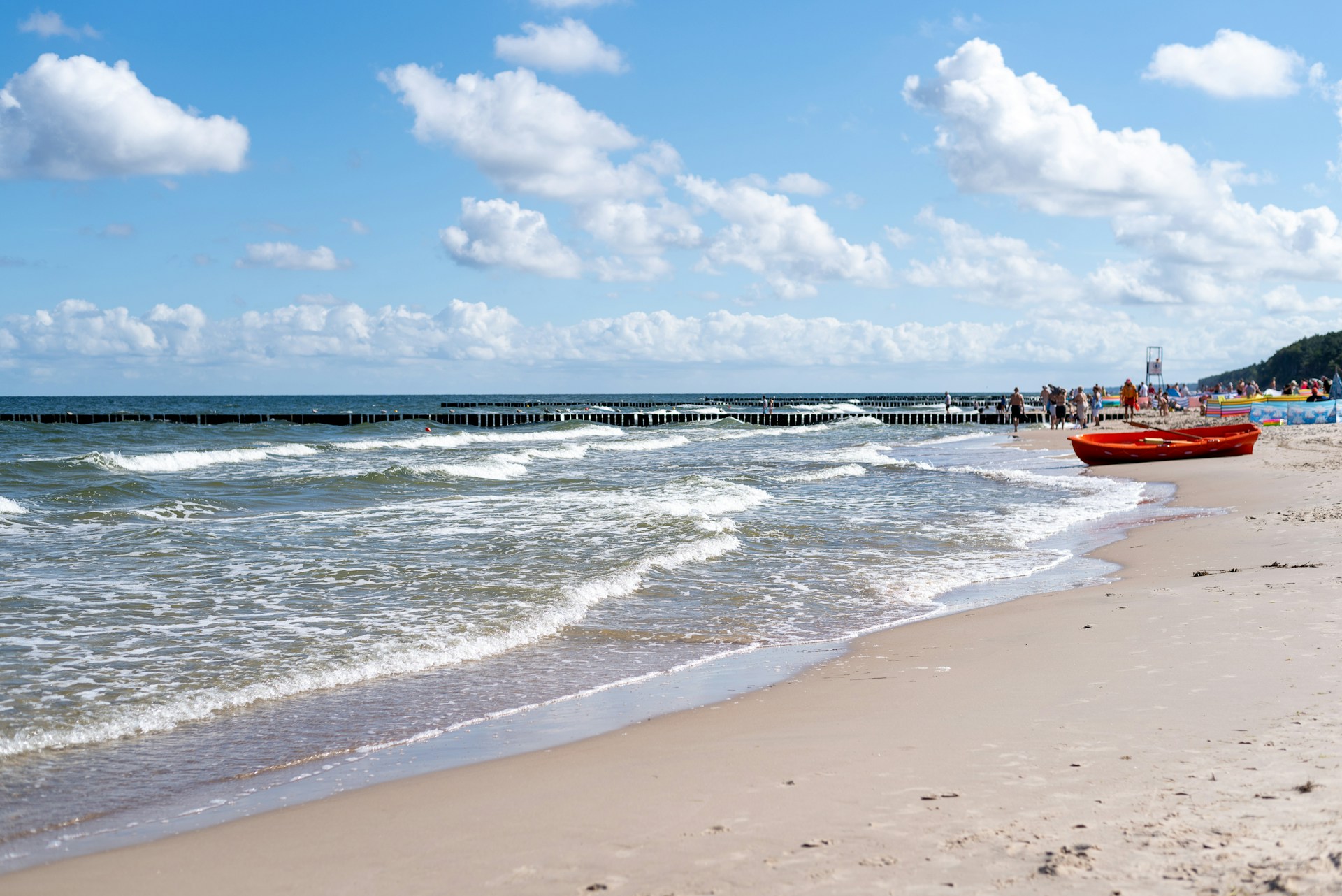 The photo presents waves washing onto a sandy beach, with a red boat and beachgoers under a blue sky with white clouds.