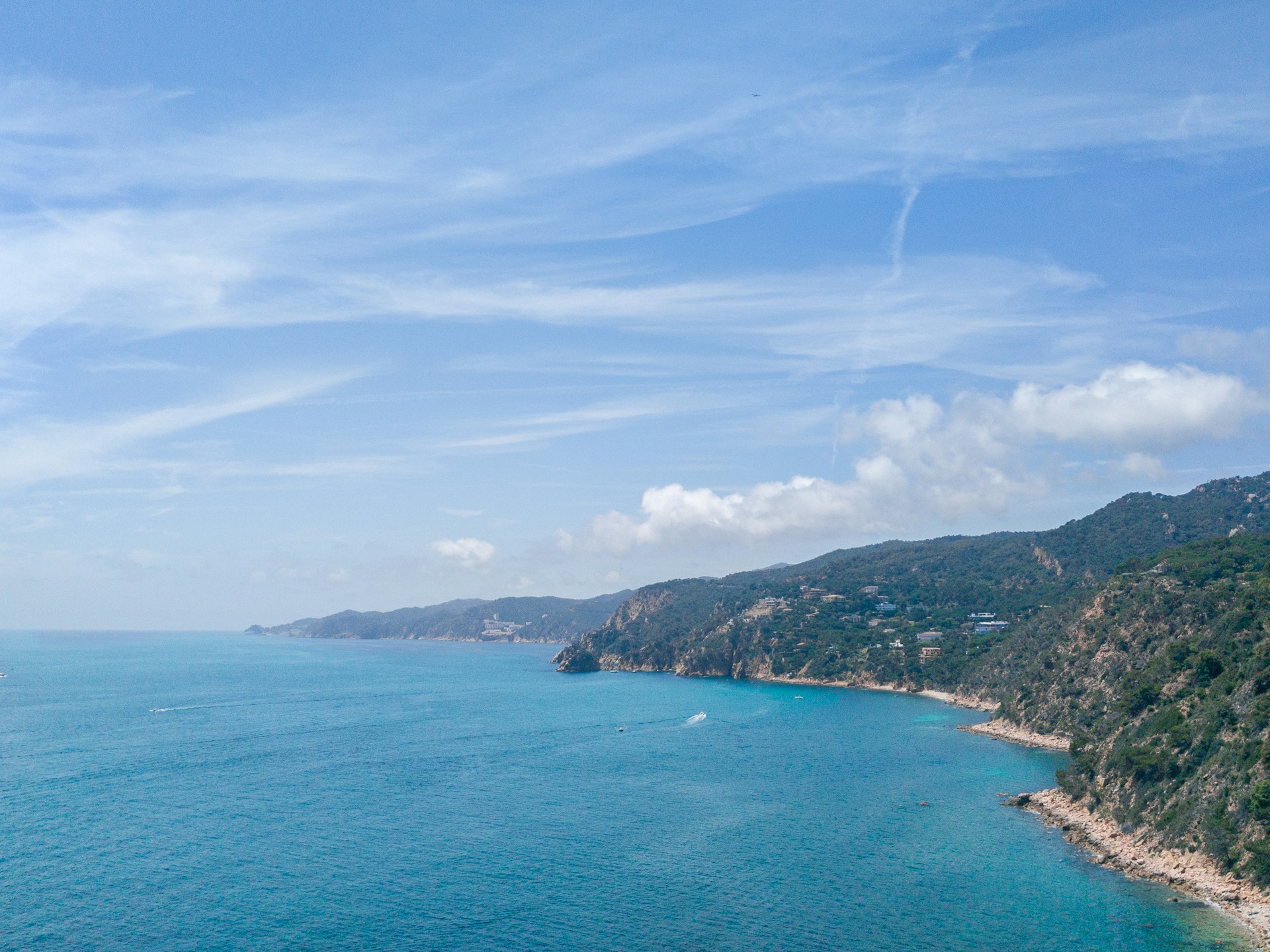 The photo presents a turquoise coastal bay bordered by rugged, forested hills with scattered houses beneath a bright, cloud-filled sky.