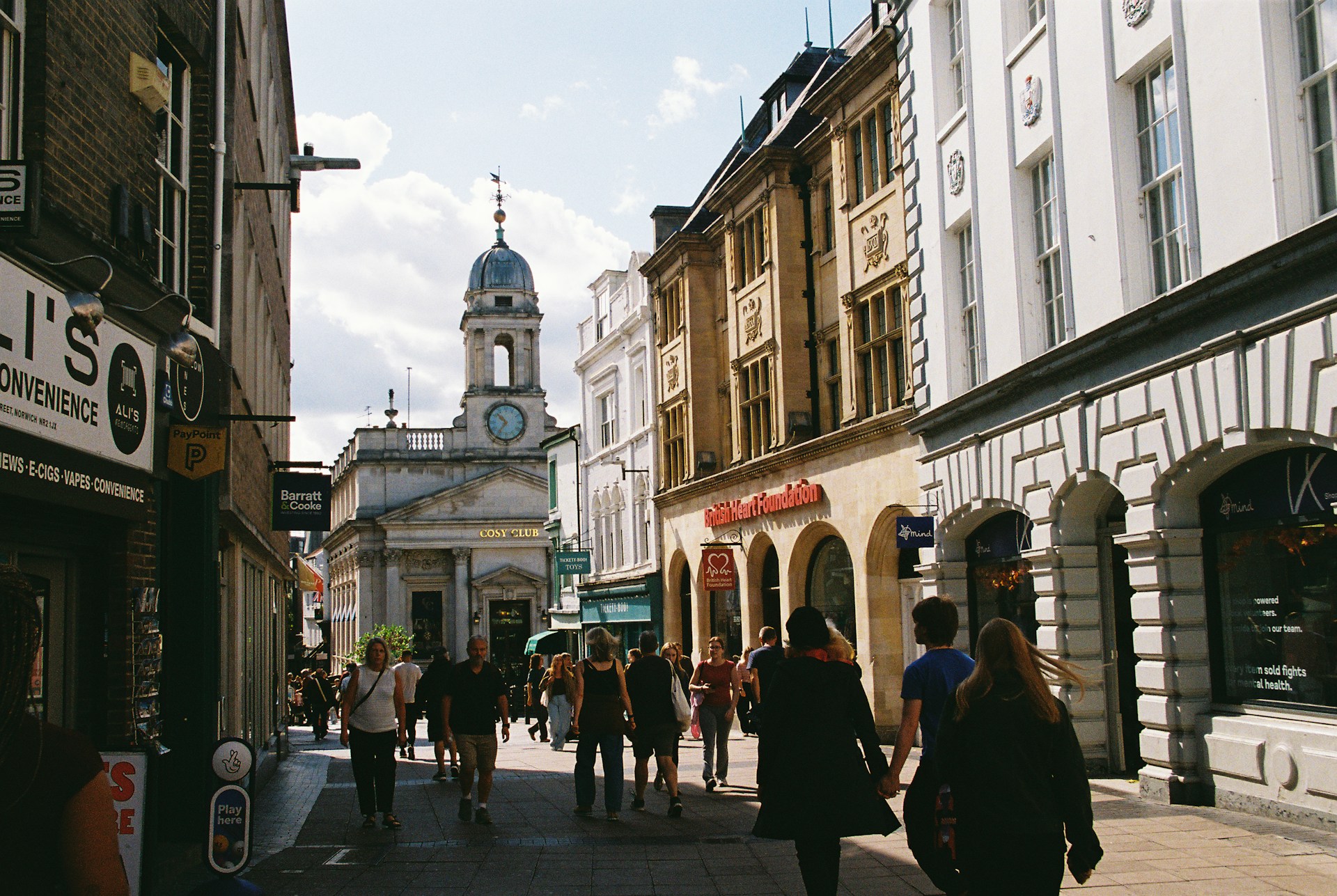 The photo presents people walking along a sunlit pedestrian shopping street lined with historic buildings, with a domed clock tower in the background.