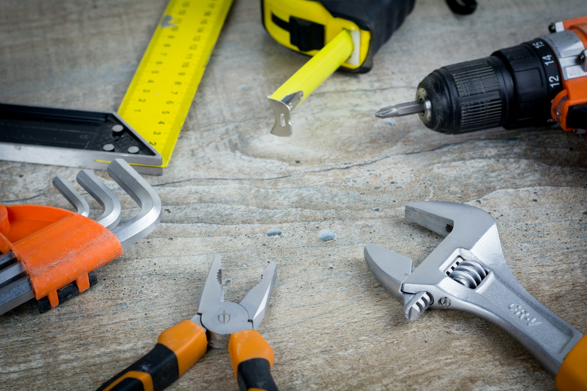 The photo presents a drill, adjustable wrench, pliers, hex keys, tape measure and carpenter’s square arranged on a wooden surface.