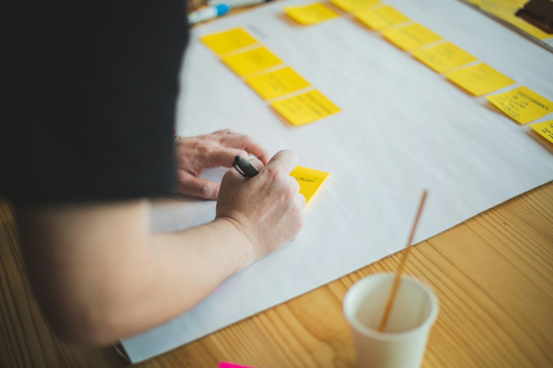 The photo presents a person writing on a yellow sticky note beside rows of notes arranged on a large sheet of paper.