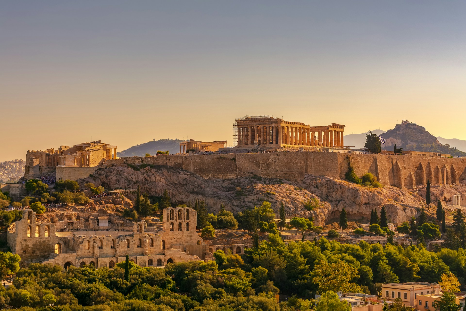 The photo presents the Acropolis of Athens glowing in warm evening light, with the Parthenon, surrounding ruins and Mount Lycabettus visible above the city.
