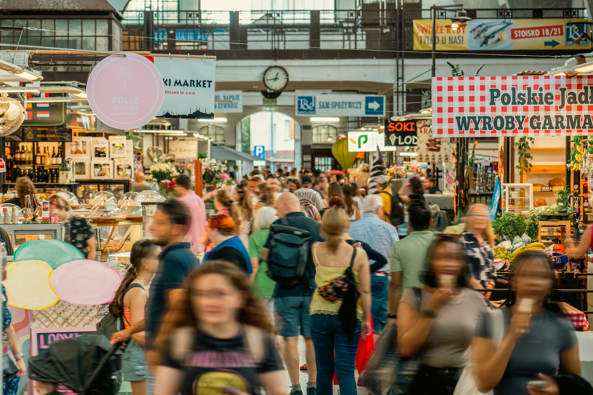 The photo presents a crowded indoor market with people walking between stalls selling food, drinks, flowers and fresh produce.