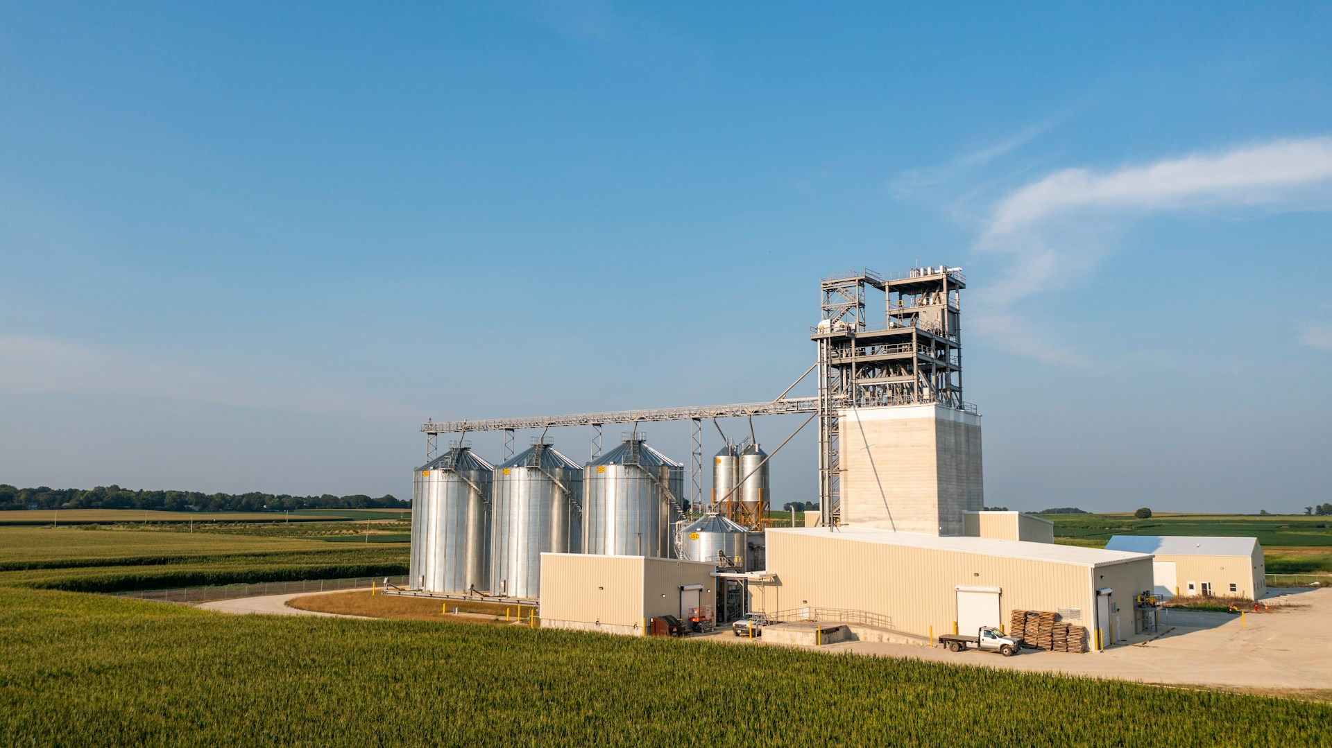The photo presents a grain processing facility with tall metal silos and a large industrial tower surrounded by agricultural fields.