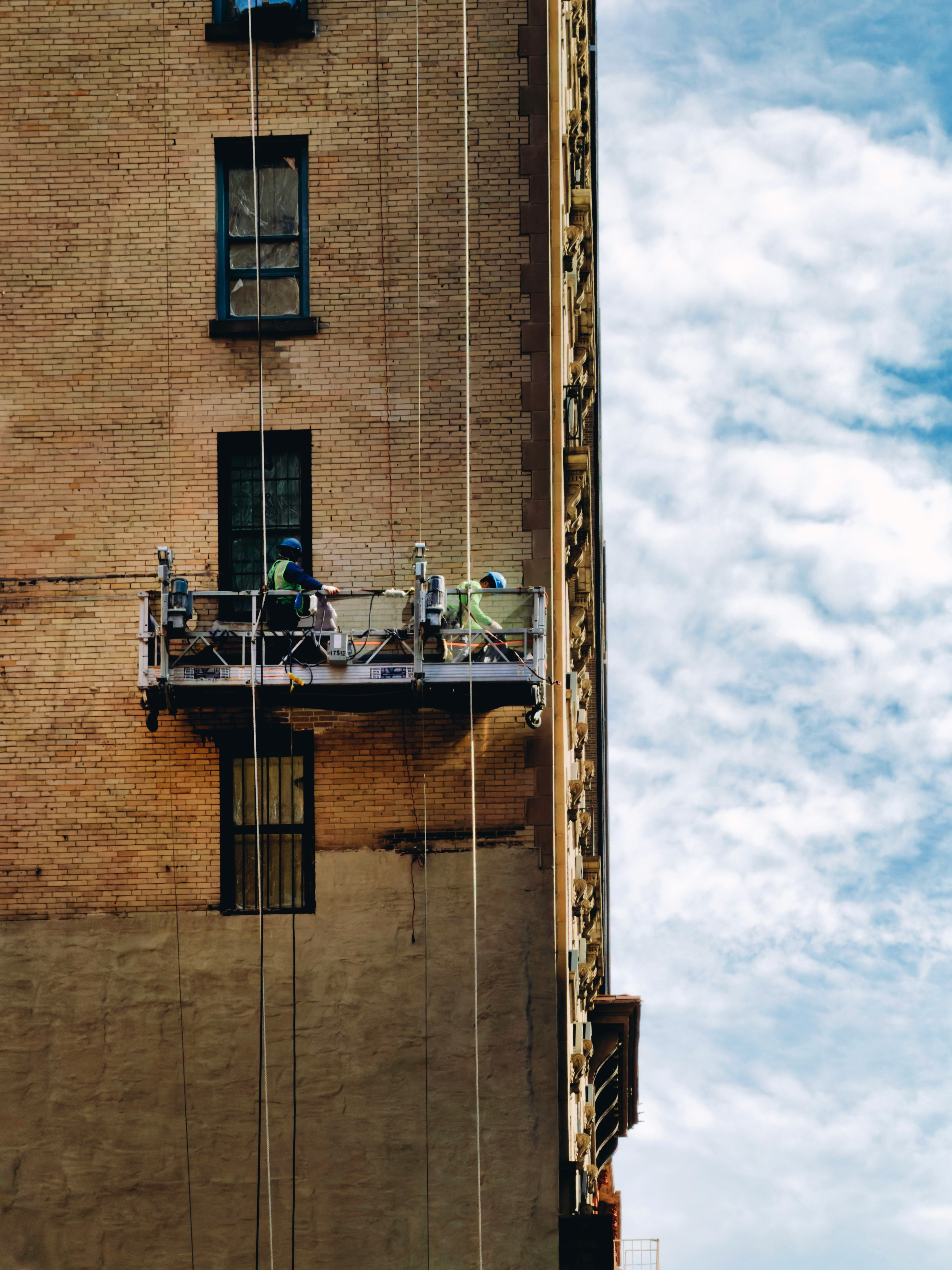 The picture shows the side of a tall brick building with two workers suspended on a motorized platform attached by vertical cables.