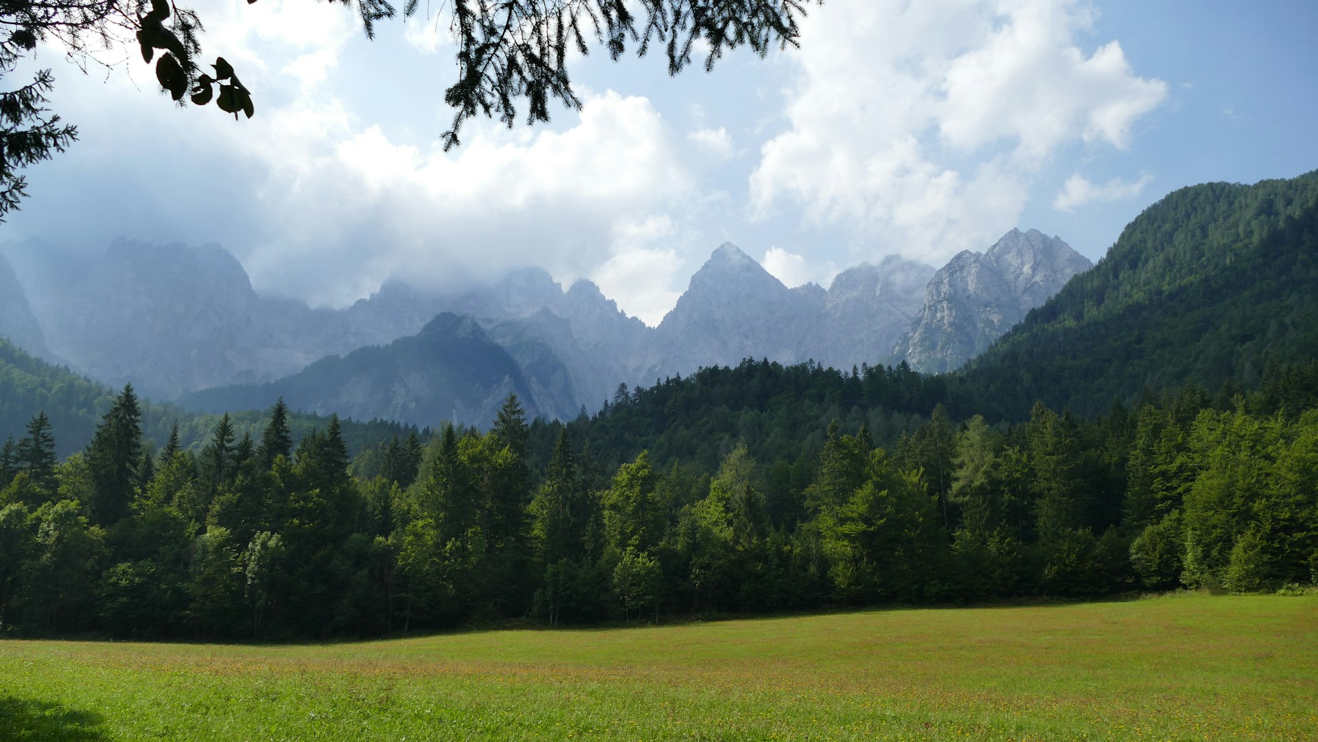The photo presents a green meadow and dense forest beneath rocky mountain peaks partly covered by clouds.