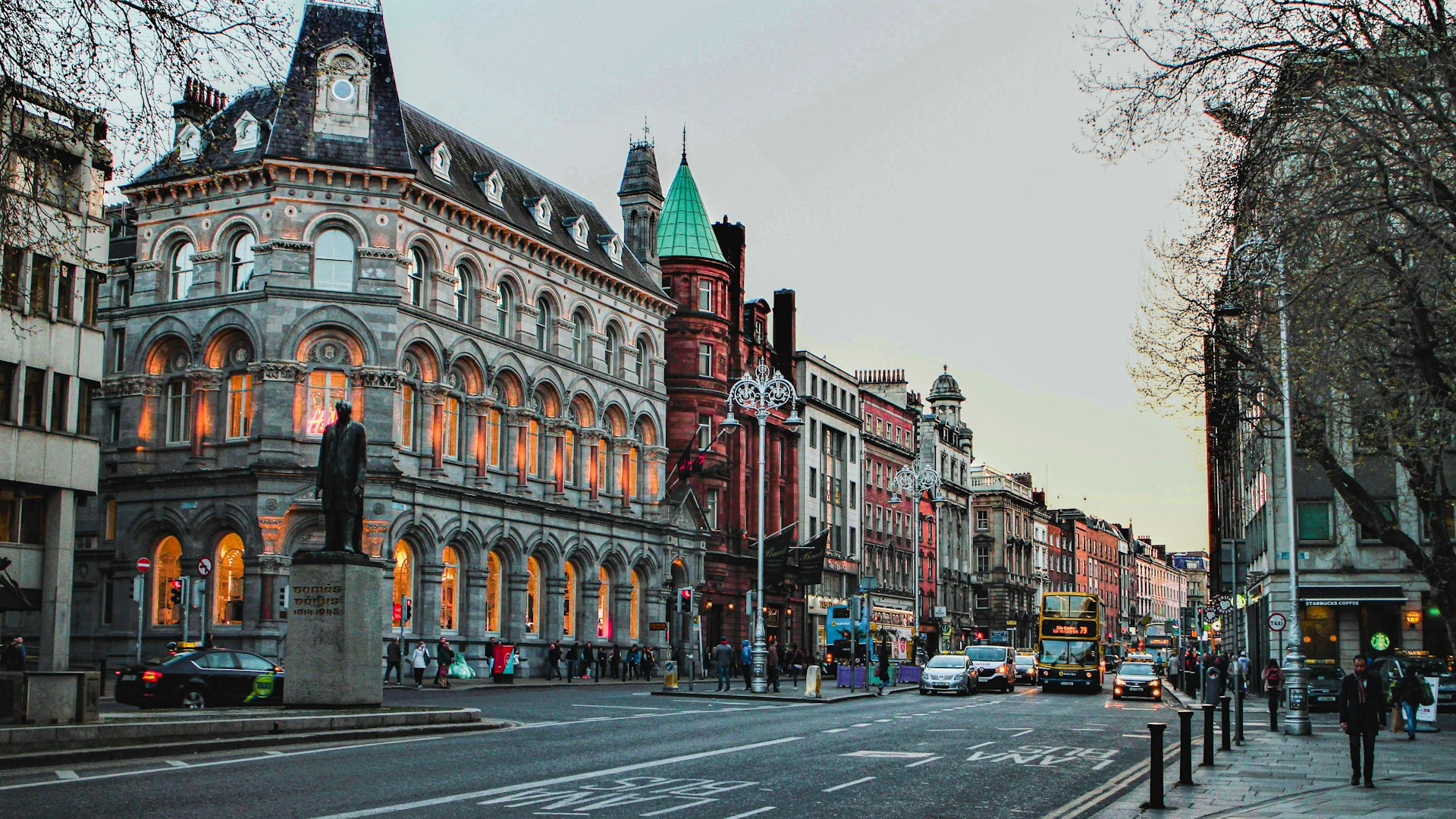 The photo presents a Dublin street lined with historic buildings, with double-decker buses, cars, pedestrians and a statue in the foreground.