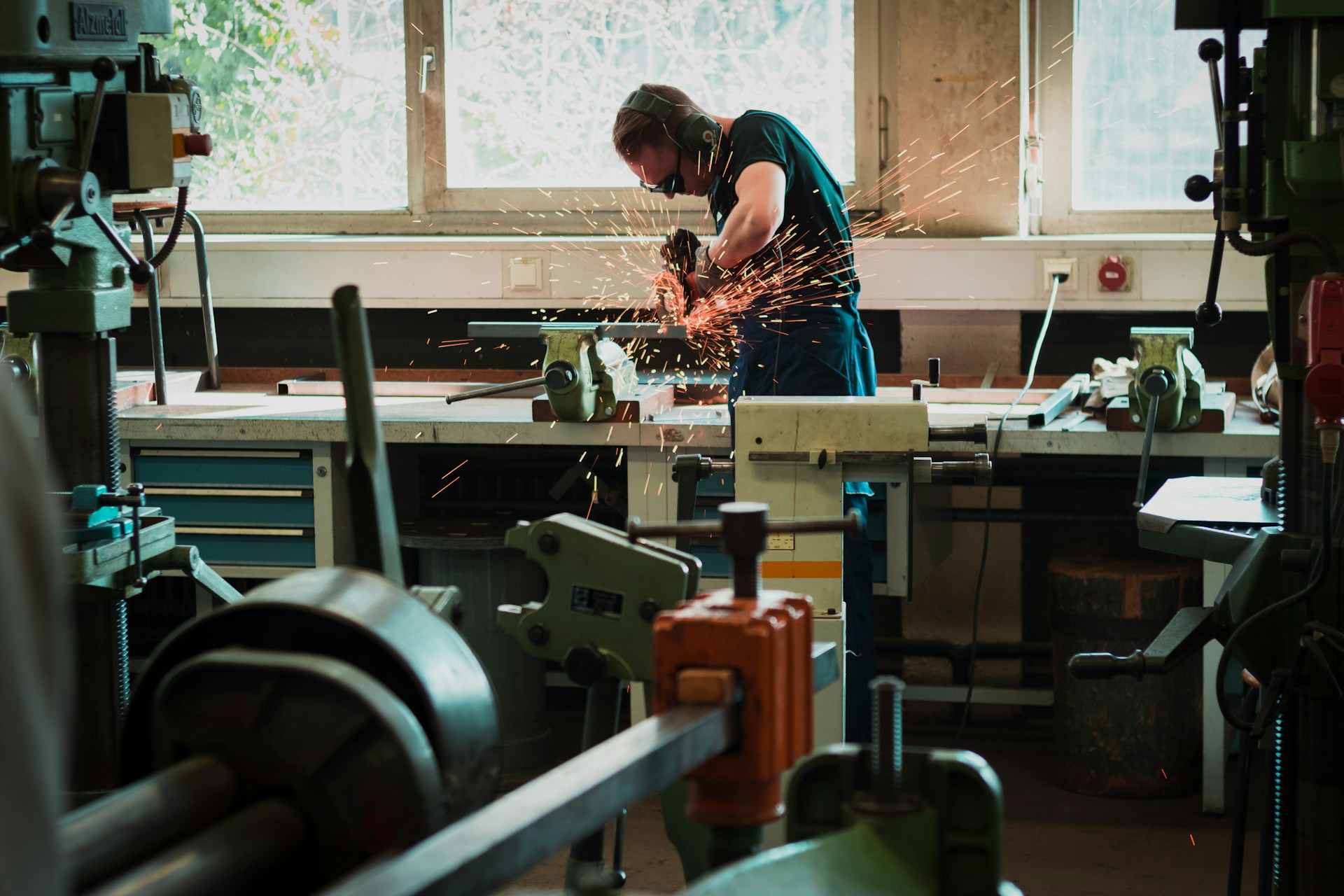 The photo presents a worker wearing safety glasses and ear protection using an angle grinder on metal, producing sparks in a workshop.
