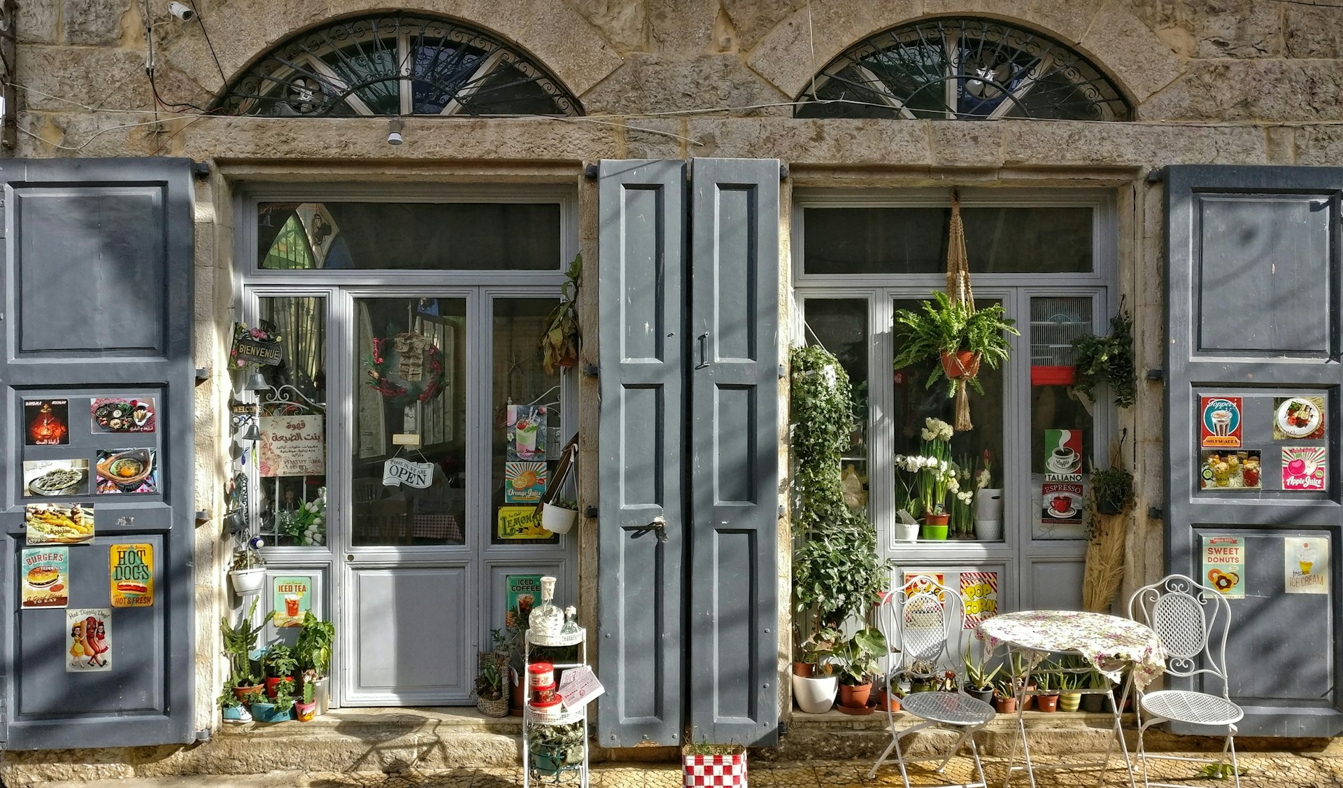 The photo presents a charming café storefront in a stone building, with grey shutters, potted plants, food posters and a small outdoor table with chairs.