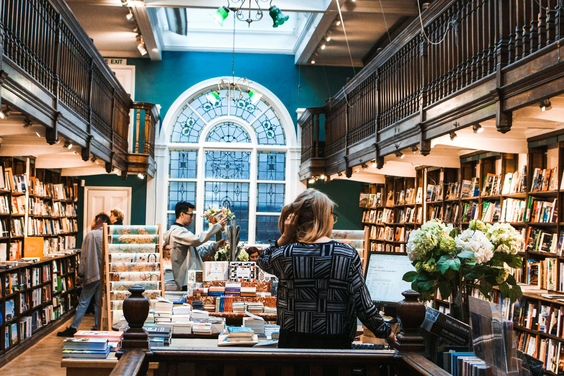 The photo presents a bookshop with wooden galleries, an arched stained-glass window, people browsing and a counter filled with books and flowers.