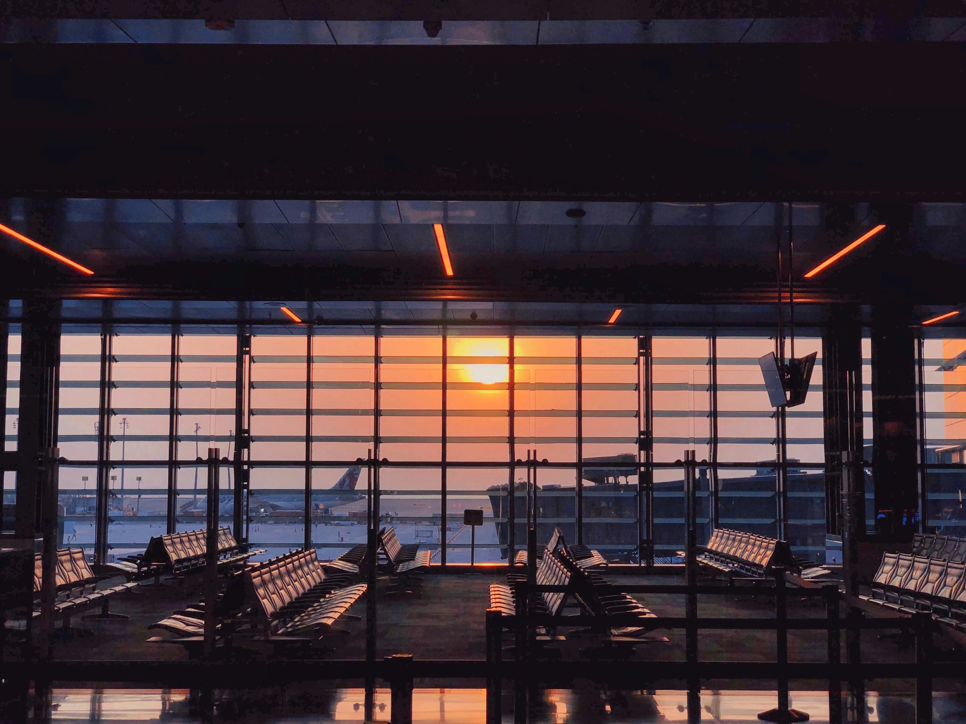 The photo presents an empty airport departure lounge with rows of seats and large windows overlooking an aircraft and an orange sunset.