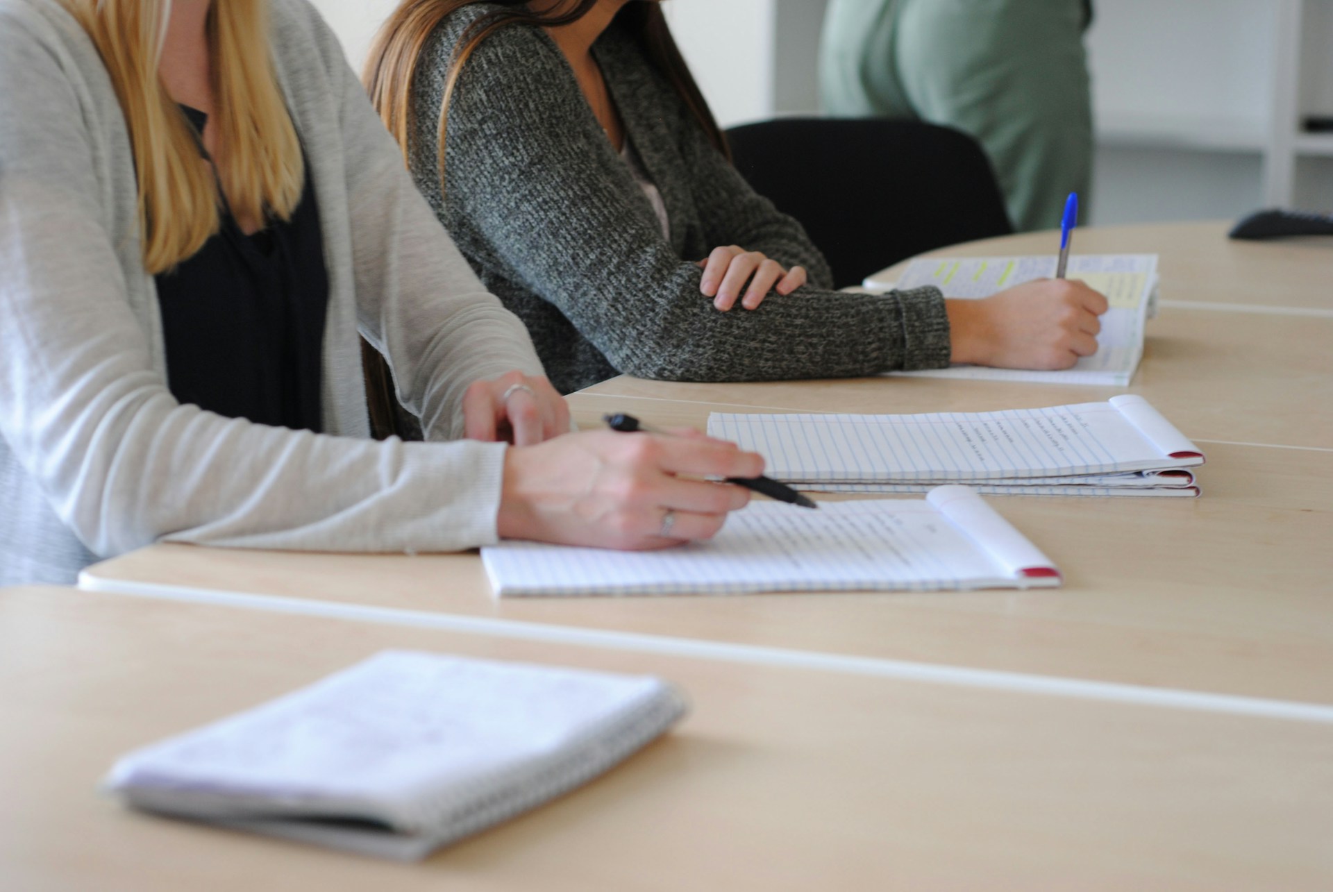 The photo presents two people seated at a table, taking notes with pens in open notebooks.