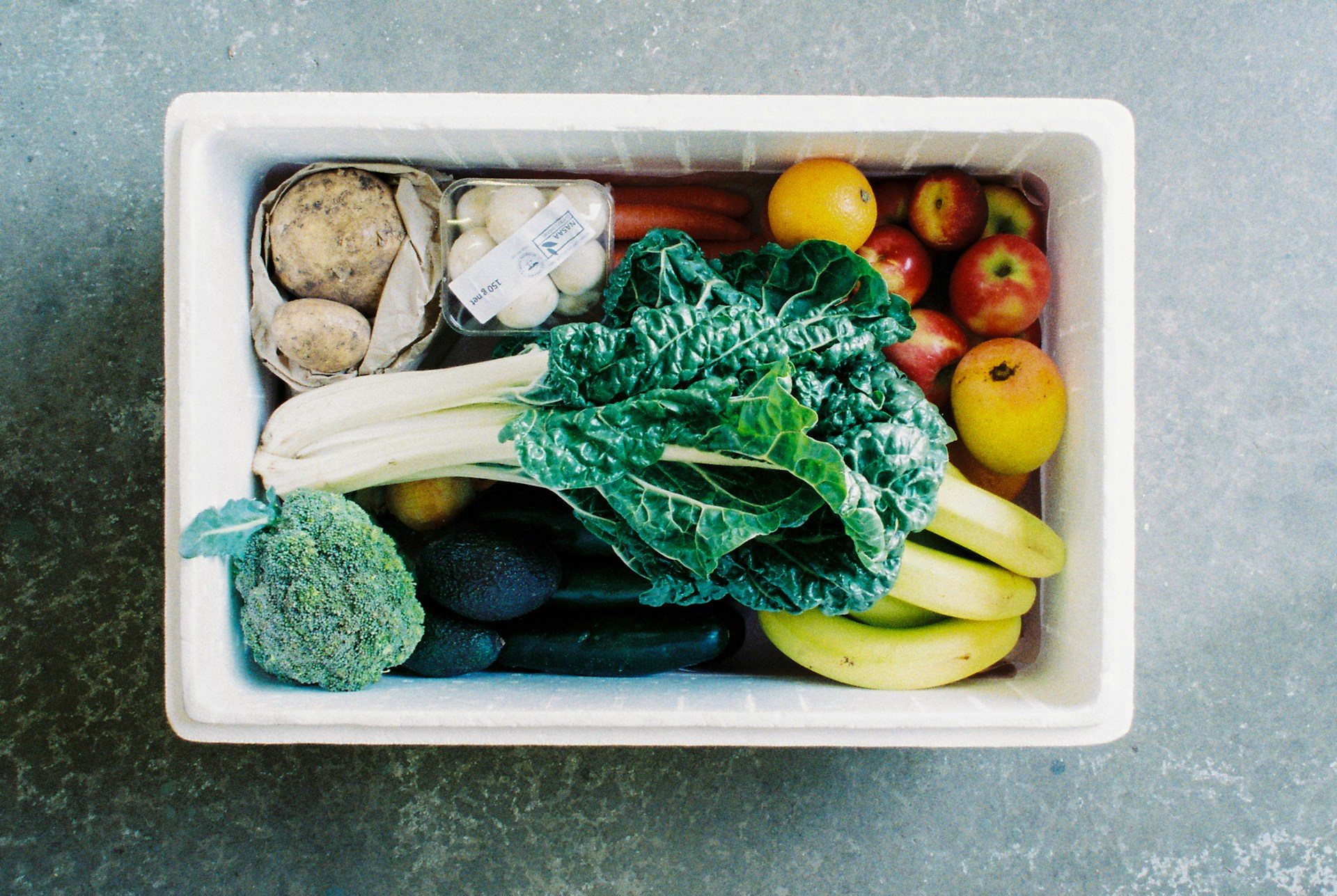 The photo presents a box filled with fresh fruit and vegetables, including leafy greens, broccoli, potatoes, carrots, apples, oranges and bananas.