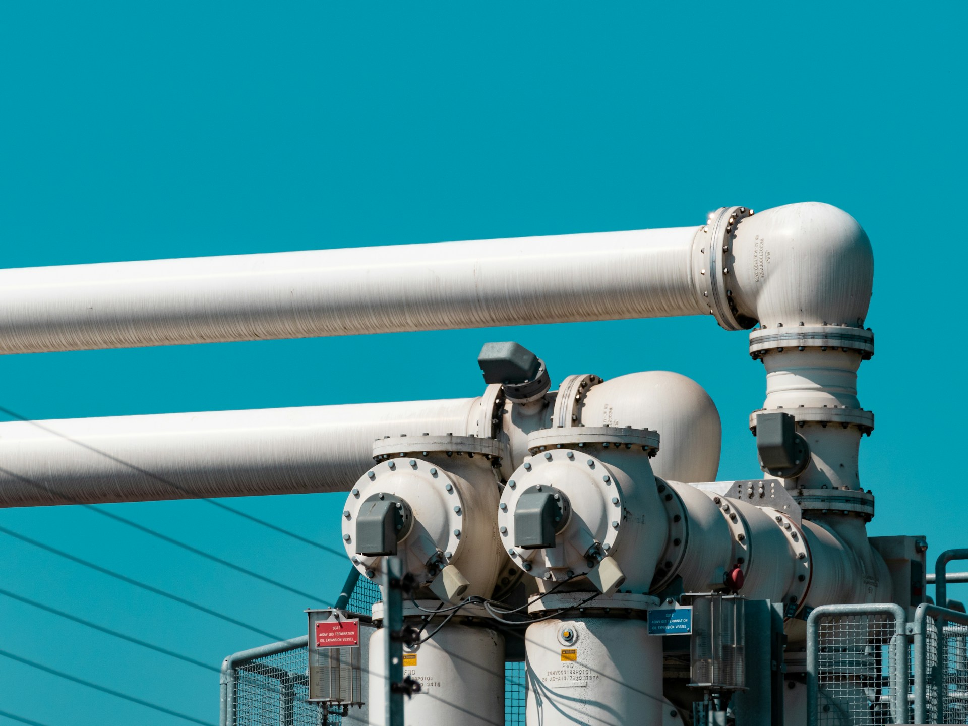 The photo presents large white industrial pipes and valves against a vivid turquoise-blue sky.