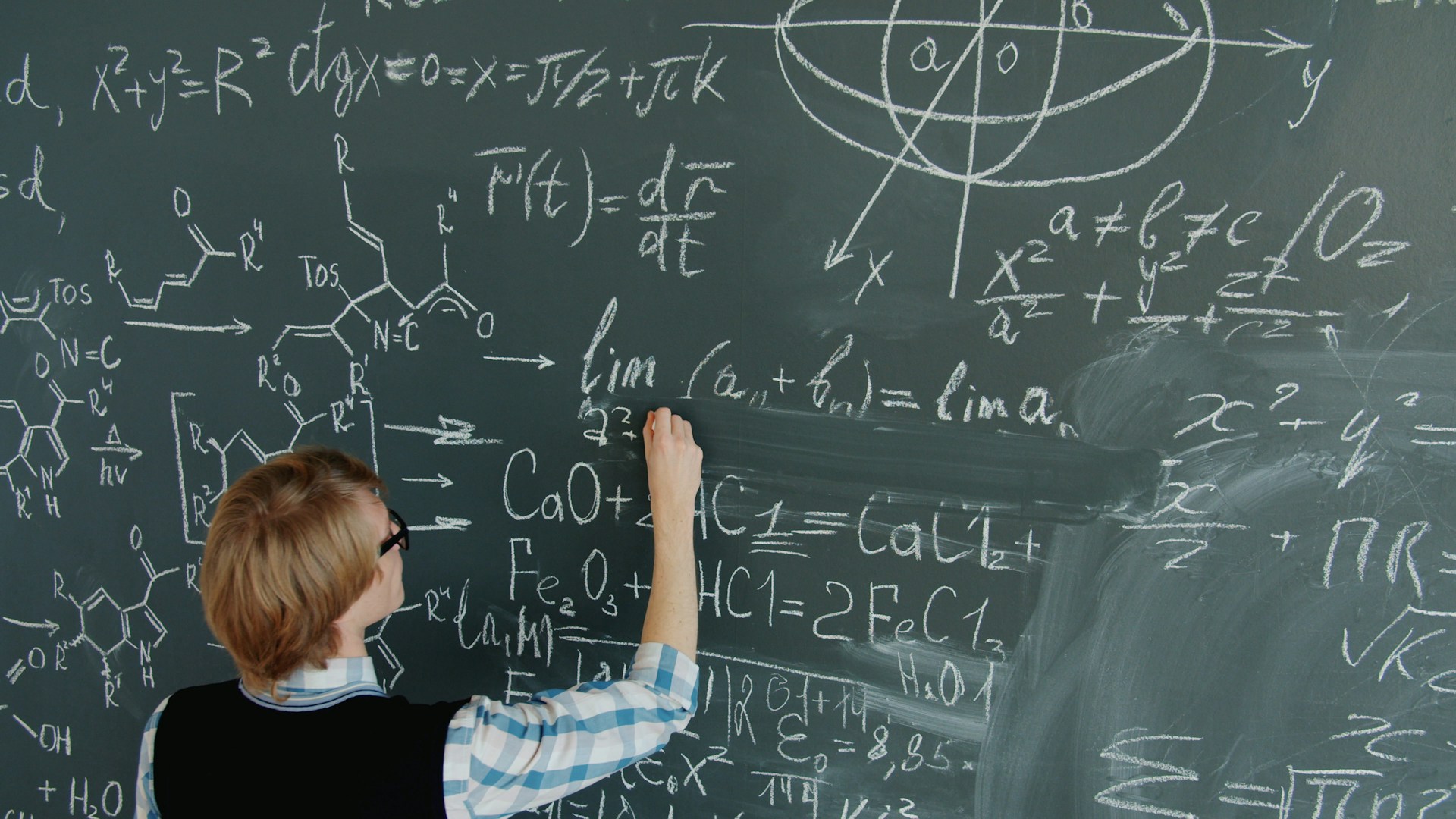 The photo presents a person writing mathematical and chemical equations on a large chalkboard.