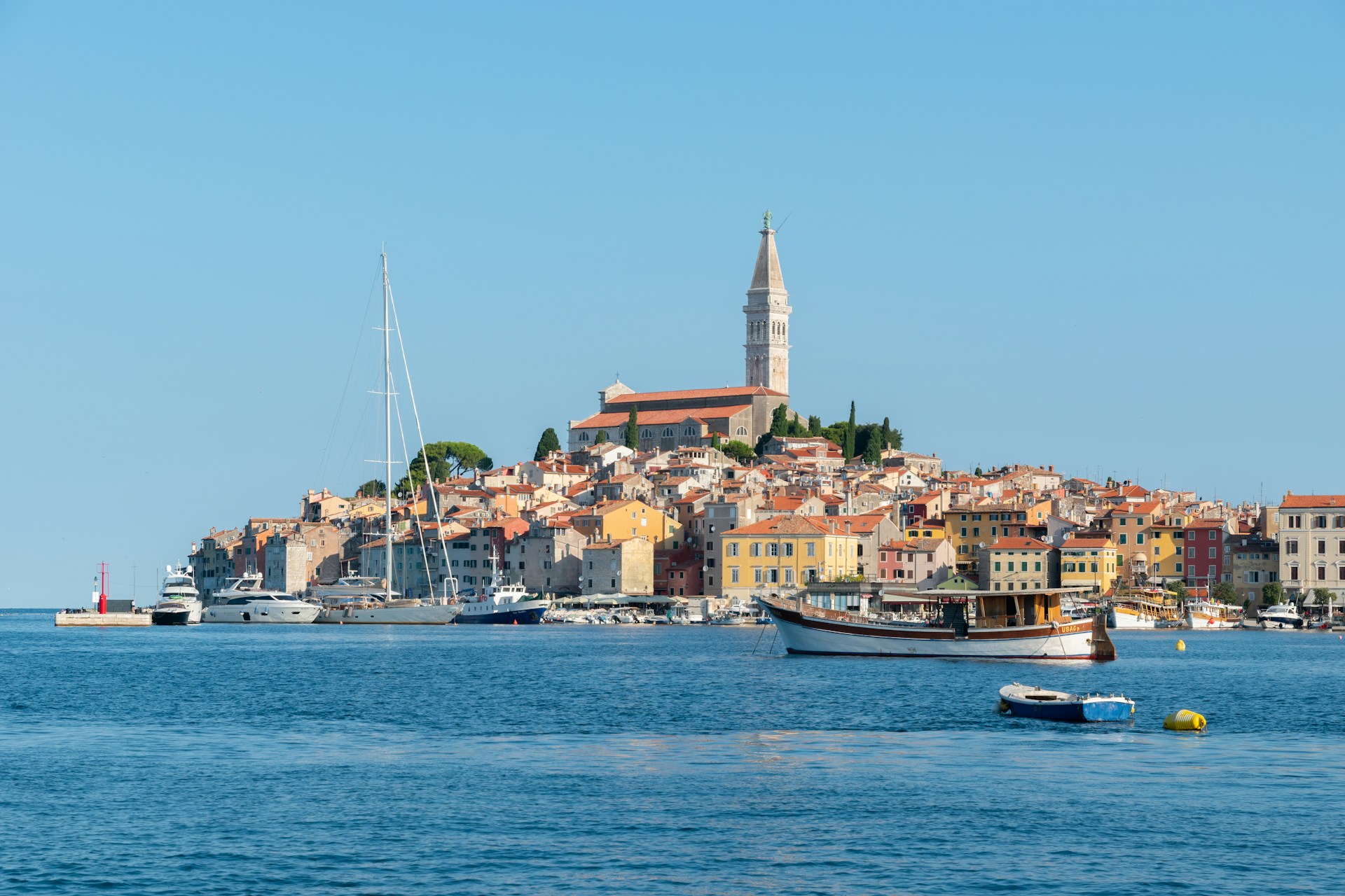 The photo presents boats in a harbour beside a coastal town with colourful buildings and a hilltop church.