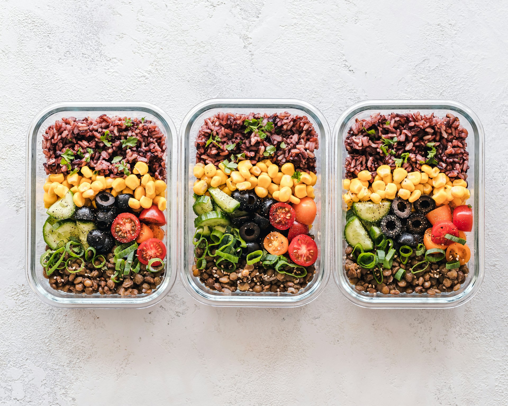 The photo presents three meal-prep containers filled with rice, lentils, corn, olives, cucumber, tomatoes, and fresh herbs.