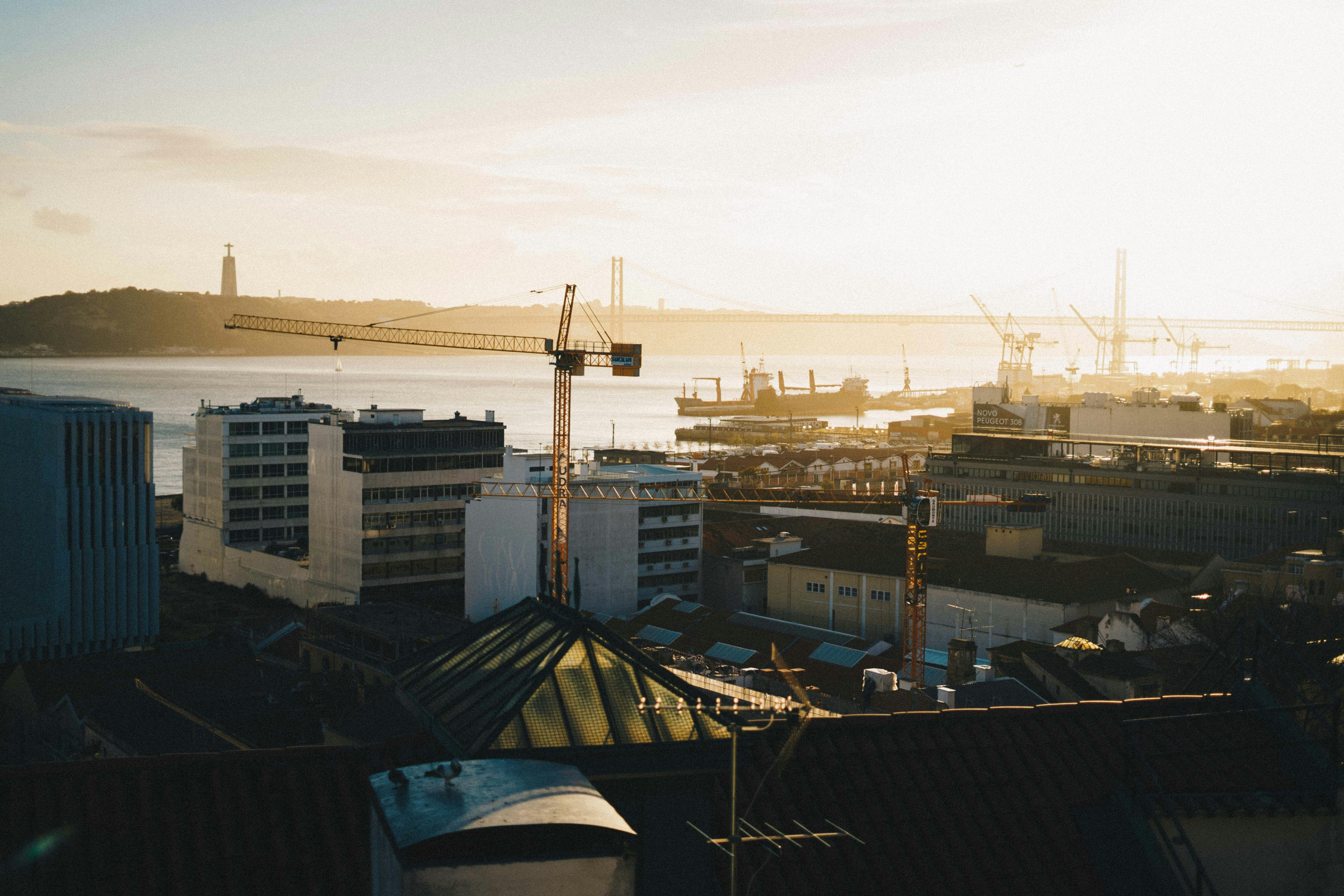 The picture shows a cityscape in Portugal overlooking a river, with industrial buildings and cranes in the foreground and a large suspension bridge in the distance.