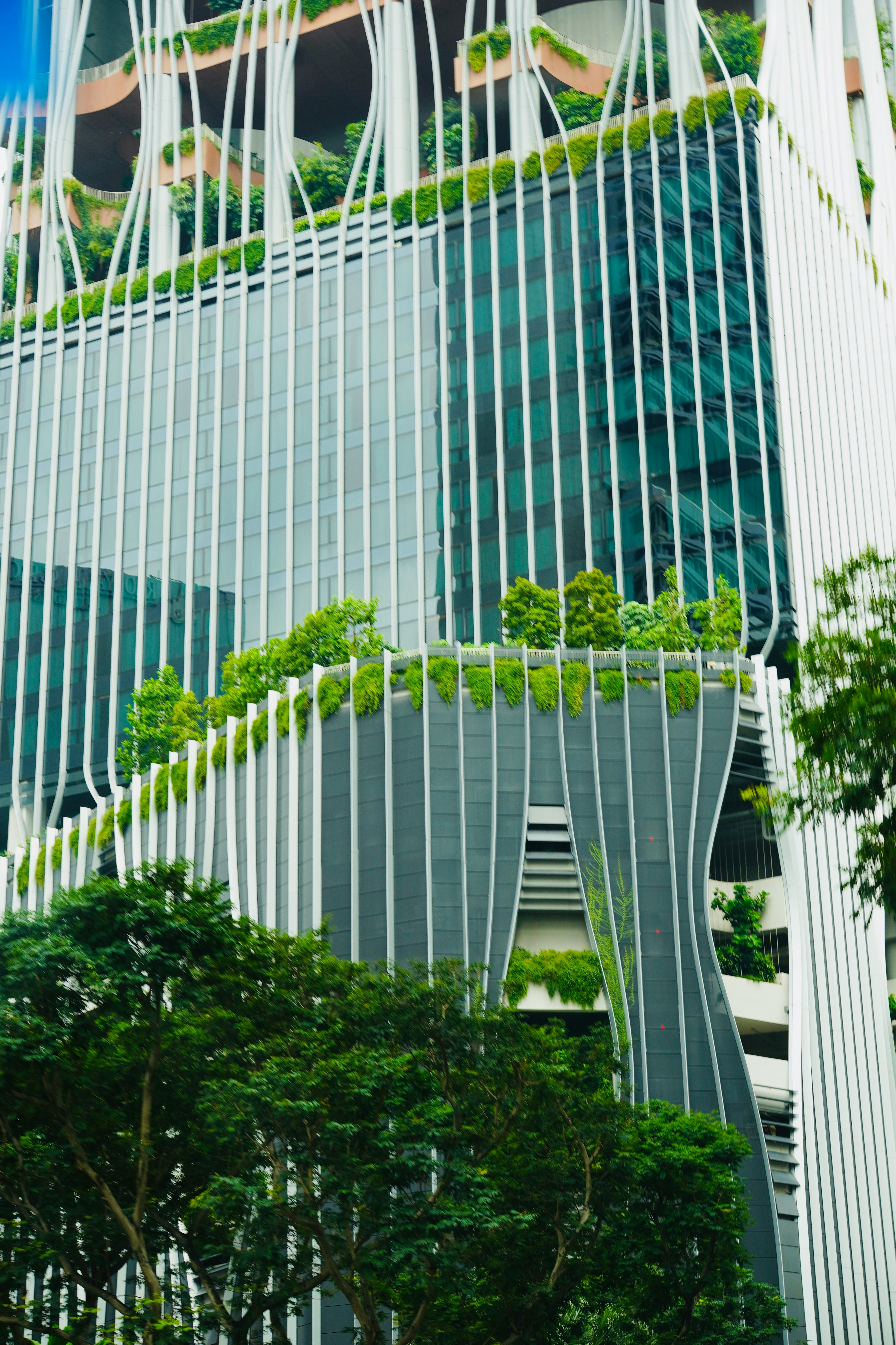 The picture shows a modern building with glass façades, distinctive white structural elements, and lush greenery integrated across terraces and balconies.