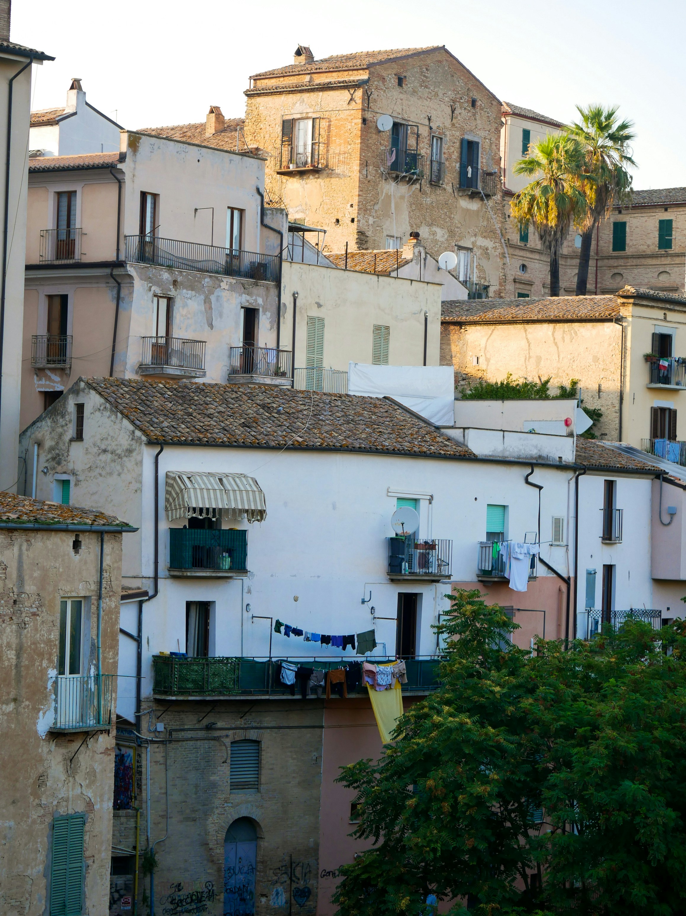 The picture shows traditional Italian buildings with tiled roofs, balconies, weathered façades, palm trees, and laundry hanging outside.