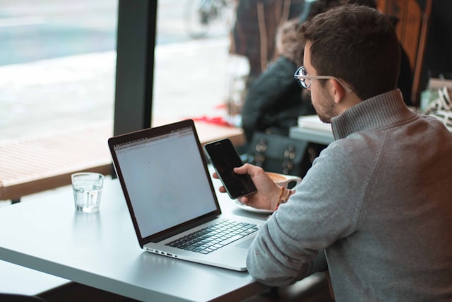 An illustrative photo of a man sitting near table with laptop and smartphone near window