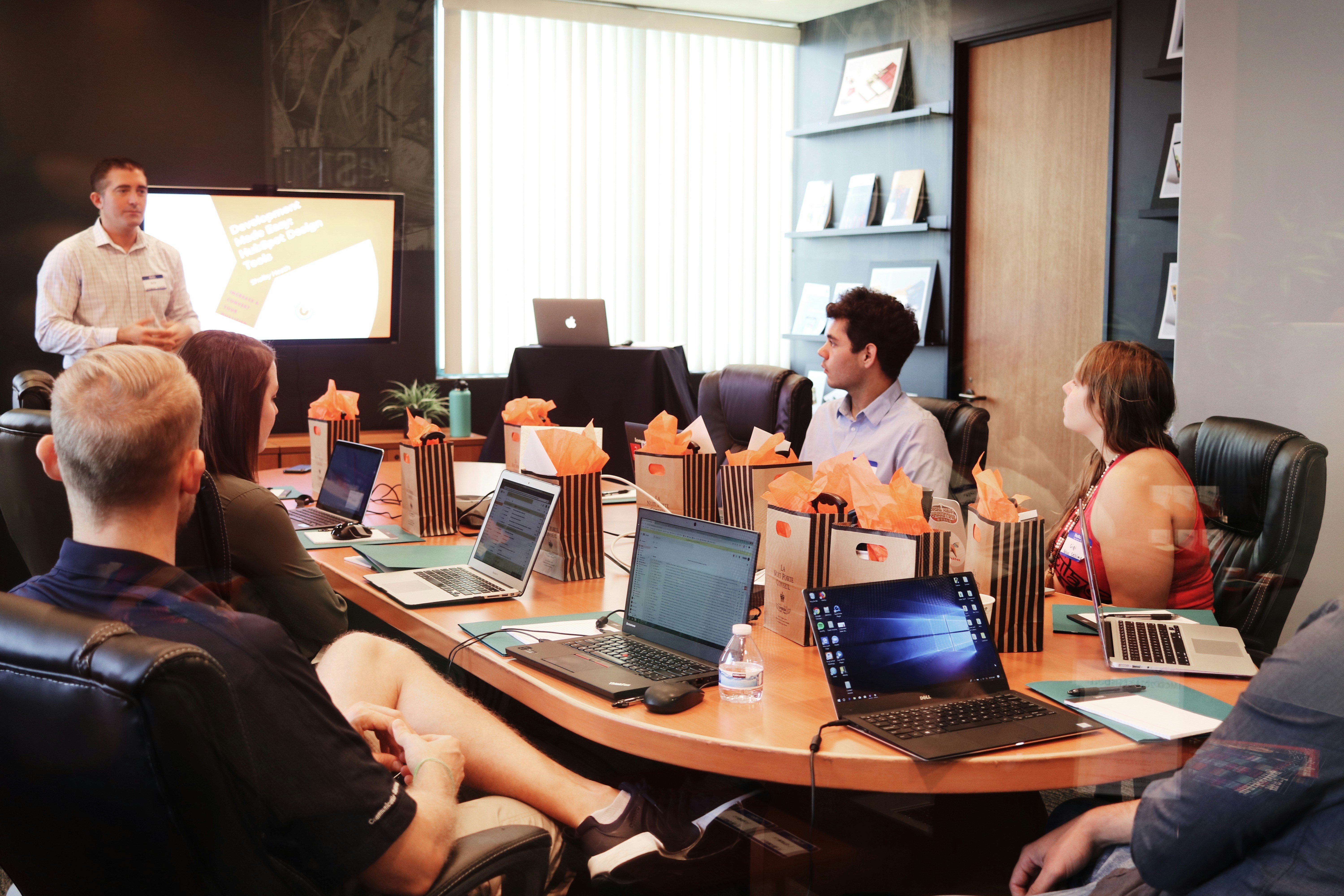 An illustrative photo of a man standing in front of people sitting beside a table with laptop computers