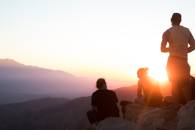 An illustrative photo of two men sitting and one man standing near a cliff during golden hour