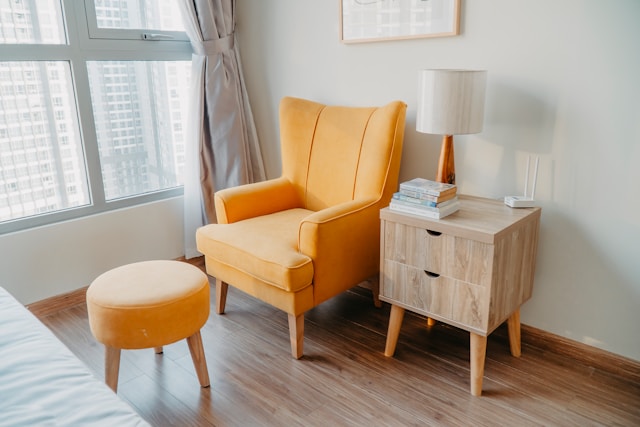 An illustrative photo of a yellow armchair and stool are placed beside a wooden nightstand by the wall