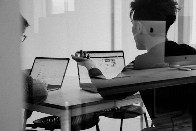 An illustrative photo of two people sitting at a desk with two laptops open in front of them