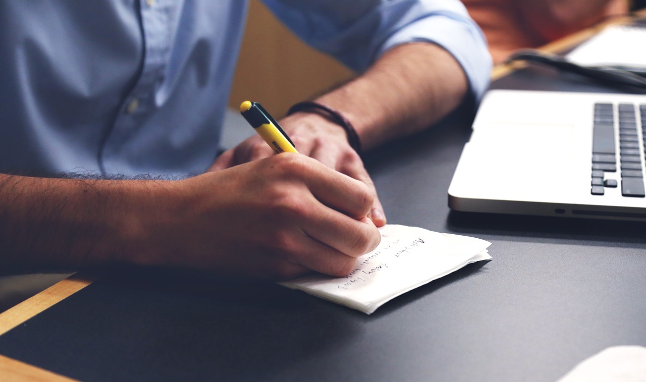 An illustrative photo of a man writing notes on paper with laptop in the background