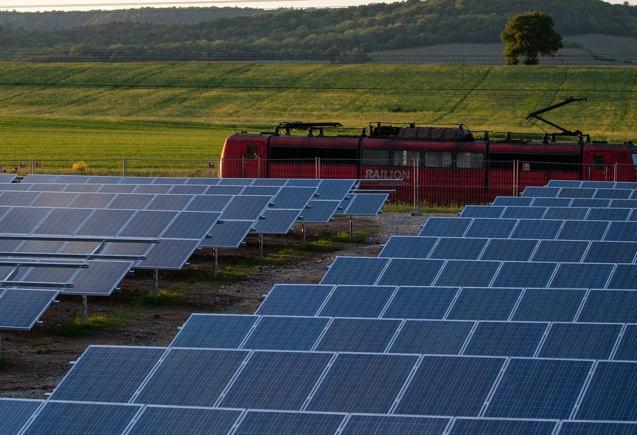 An illustrative photo of a field of solar panels in the foreground with a red electric train passing by in the background