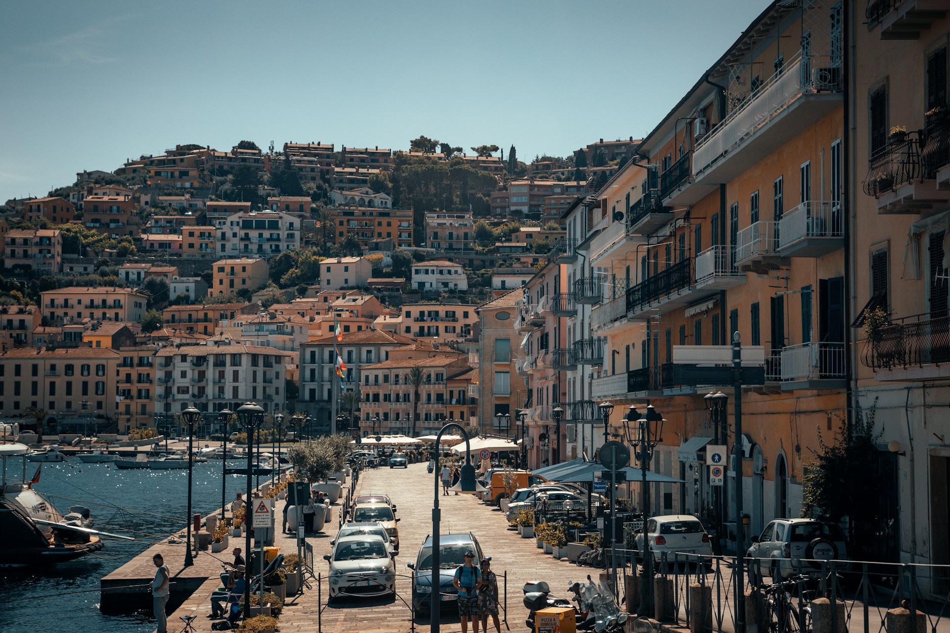 An illustrative photo of vehicles parked on the coast with buildings near a body of water