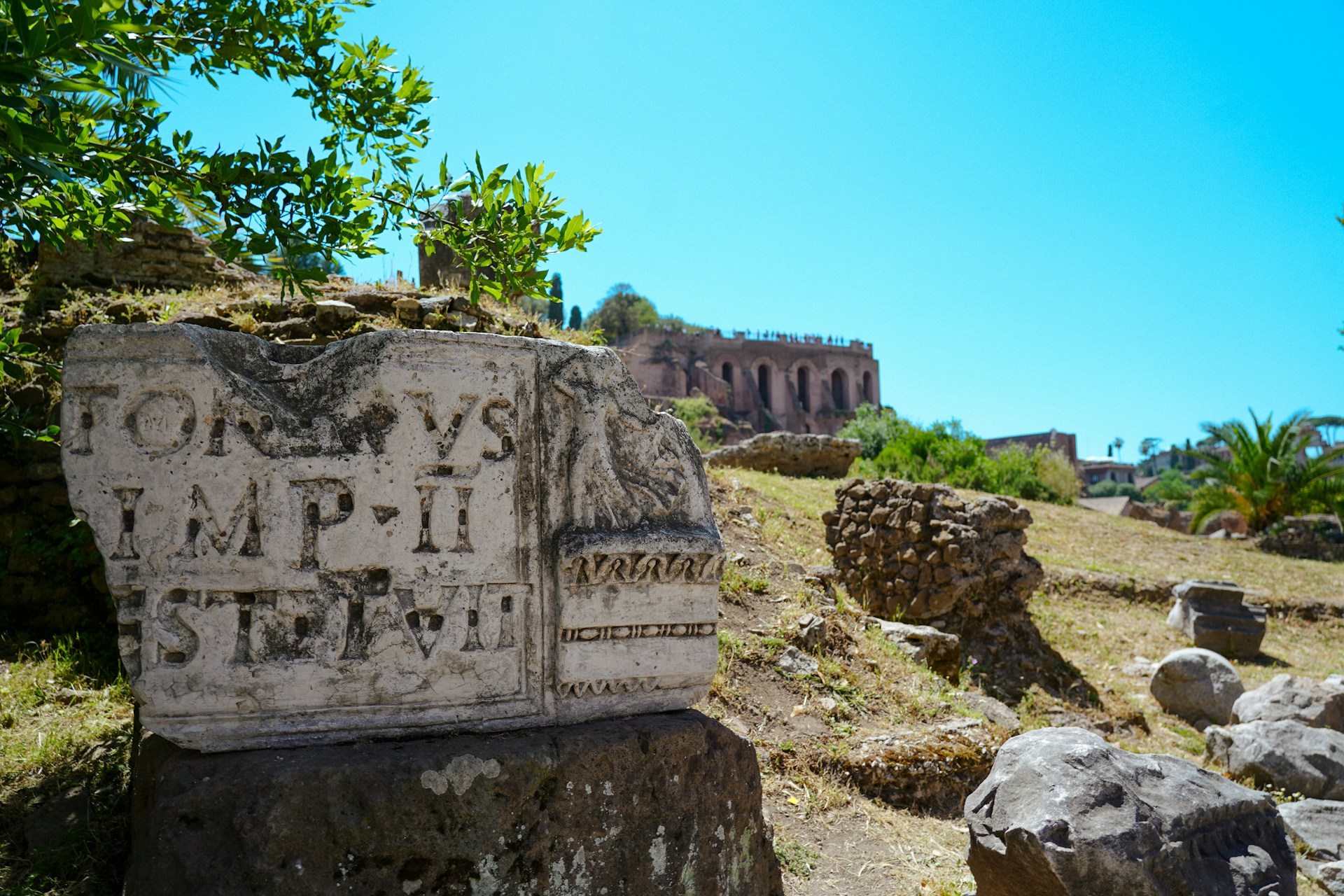 An illustrative photo of an ancient stone inscription with Latin text, set against a backdrop of greenery and ruins under a clear blue sky