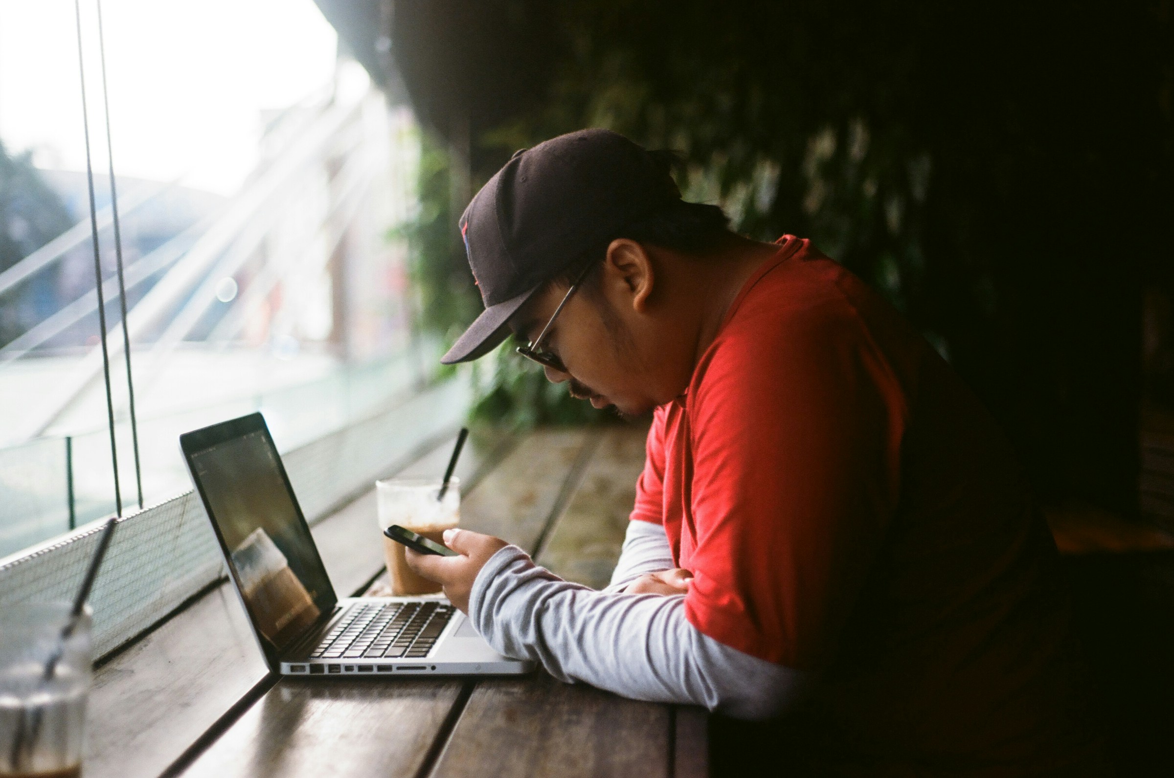 An illustrative photo of a person sitting at a wooden table, using a laptop and holding a mobile phone.