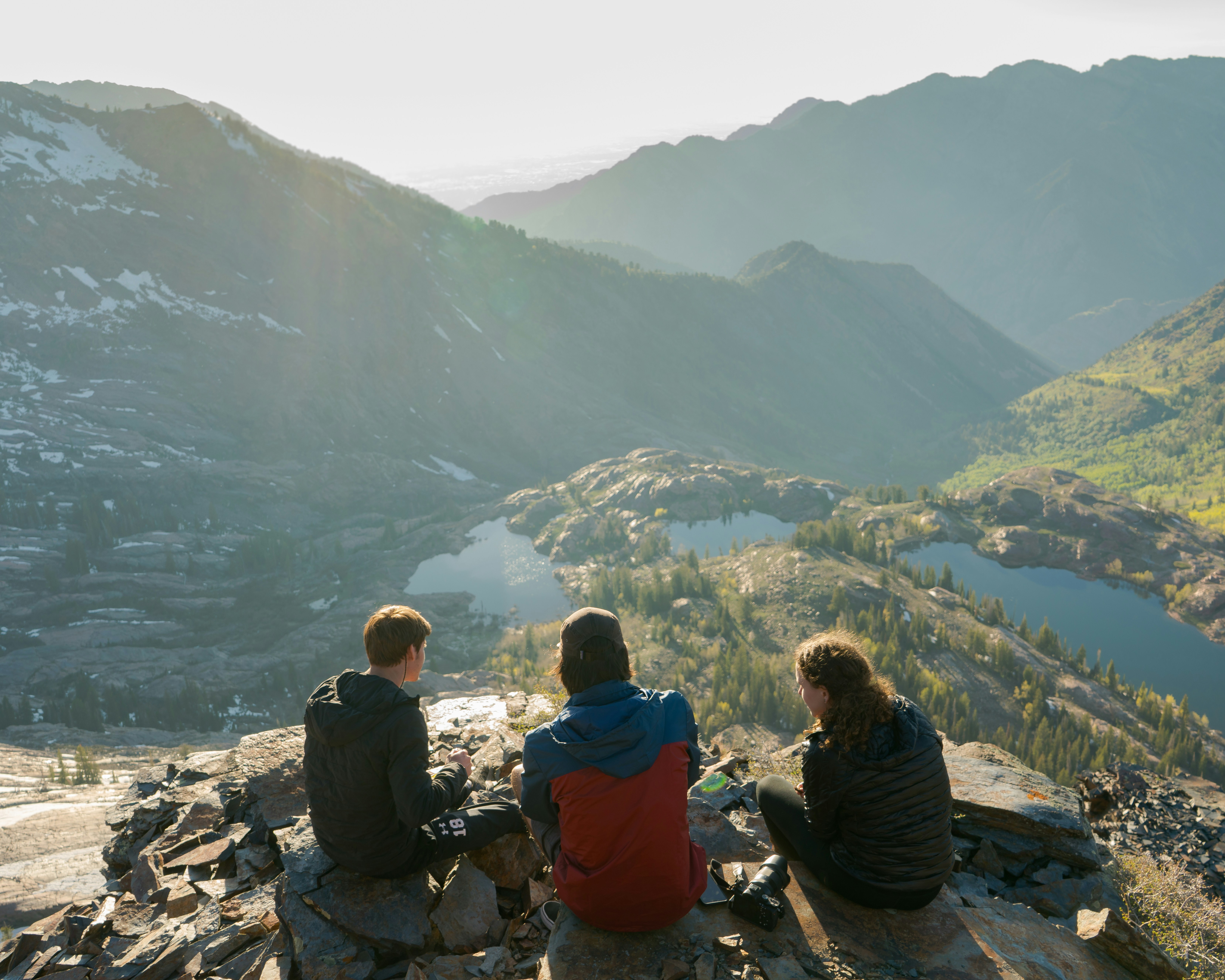 Image by Unsplash An illustrative photo of three people sitting on cliff overlooking lake and mountains during daytime.
