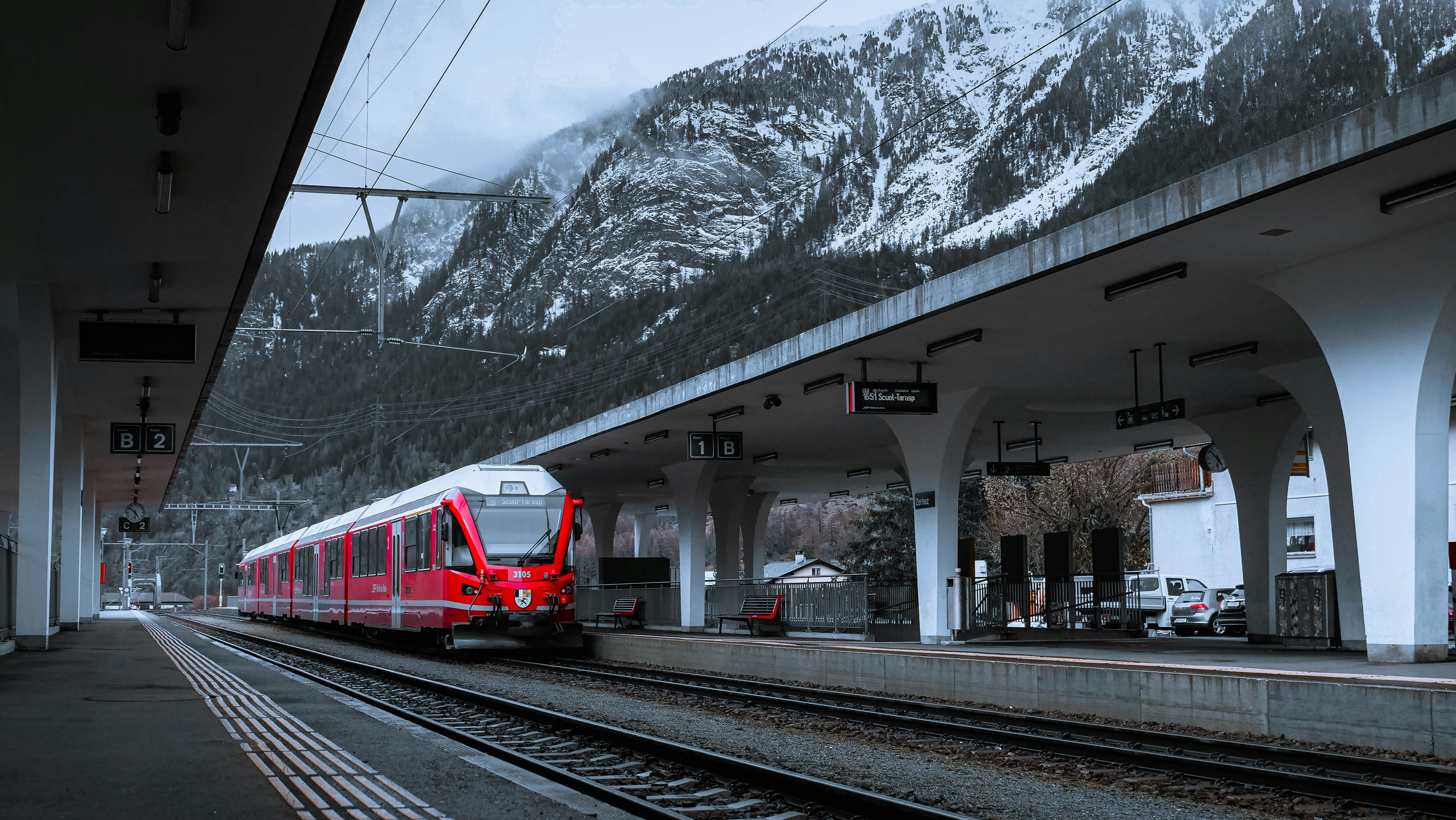 An illustrative photo of a red train traveling down train tracks next to a snow covered mountain.